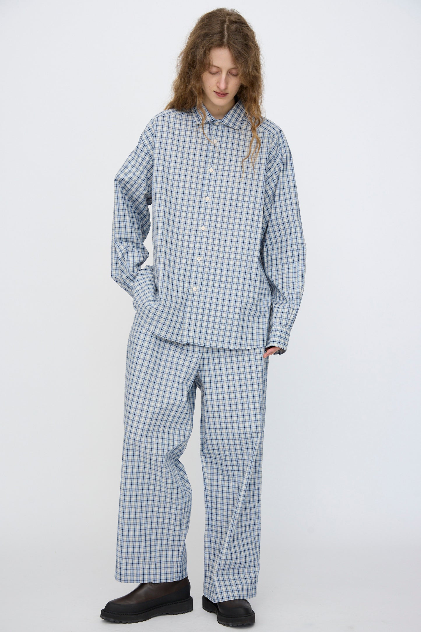 A person with long curly hair wears the Baserange Claude Shirt in Blue Check as a loose-fitting pajama set, paired with black shoes, standing against a plain white background.