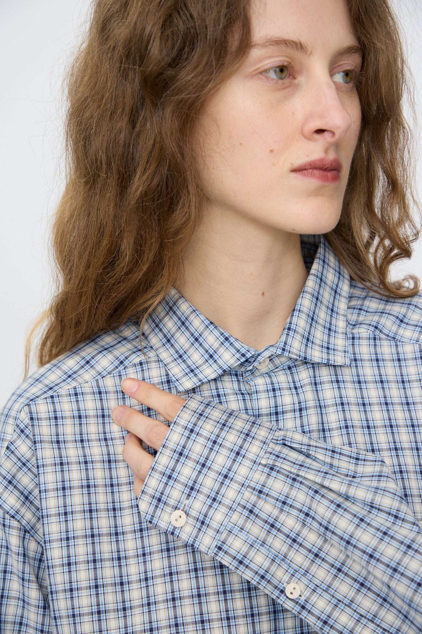 A person with long, wavy hair wears the Baserange Claude Shirt in Blue Check, holding the collar with one hand and looking off to the side.