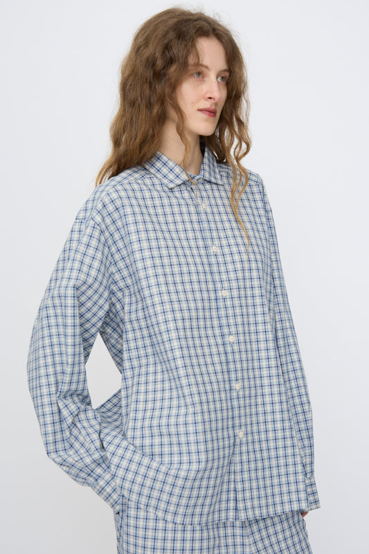 A person wears the Baserange Claude Shirt in Blue Check, a loose-fitting button-up, standing with hands in pockets against a plain background.