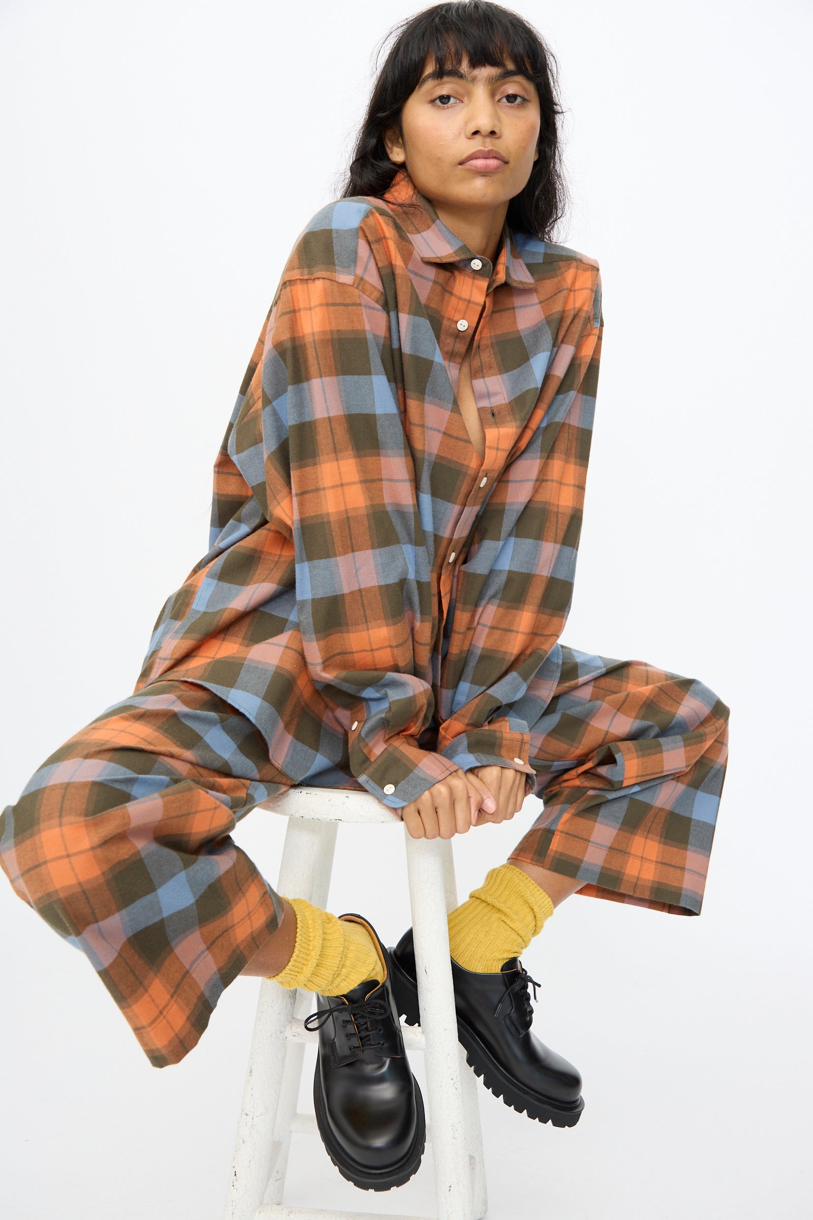 A person sits on a white stool against a plain background, wearing the Baserange Claude Shirt in Frame Plaid with yellow socks and black shoes.