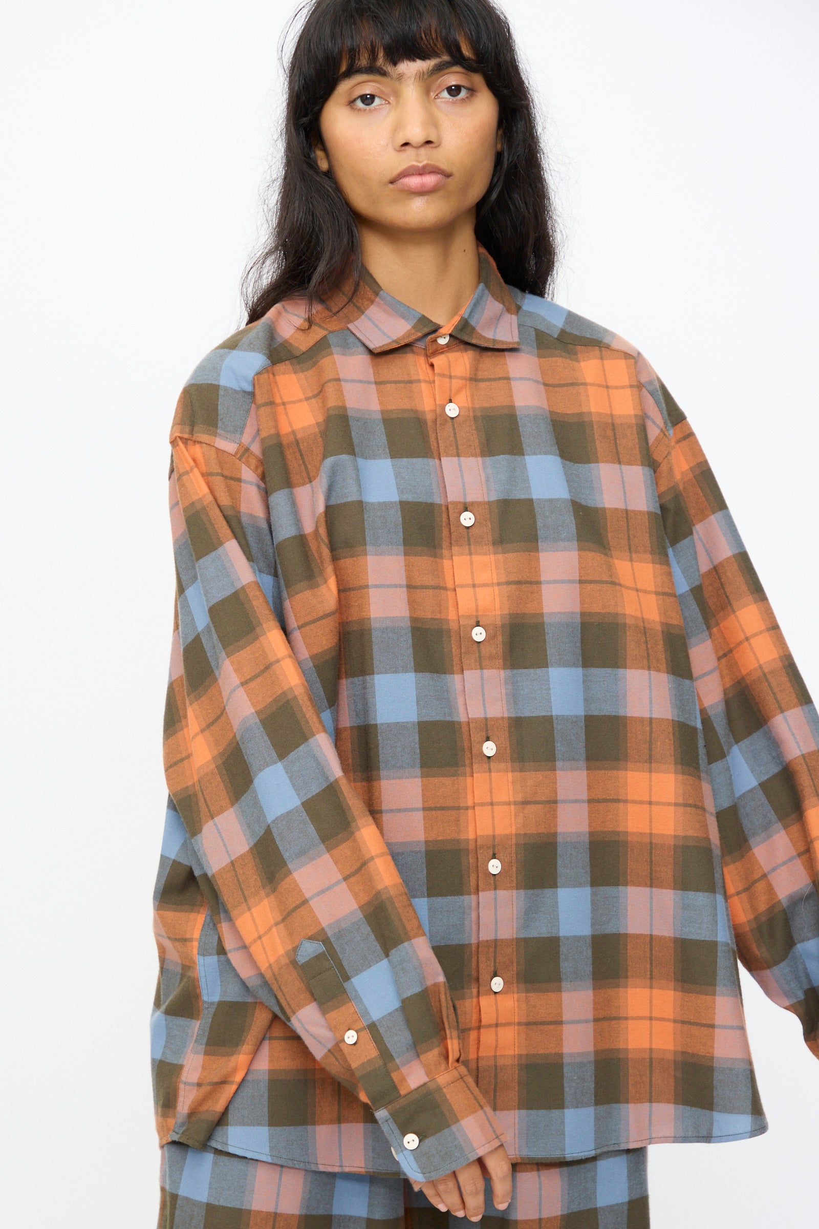 A person stands against a plain light background wearing the oversized Baserange Claude Shirt in Frame Plaid, an organic cotton button-up featuring an orange, blue, and olive plaid pattern.