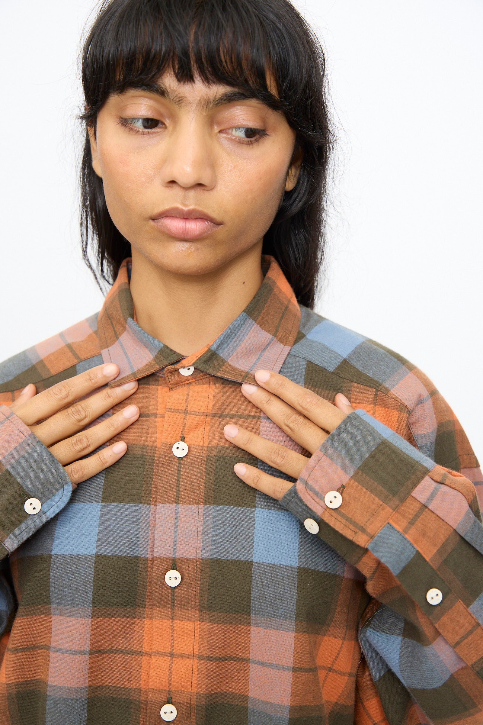 A person wears the Baserange Claude Shirt in Frame Plaid—an oversized button-up with orange, blue, and green tones, featuring buttoned cuffs—holding both hands near the collar and gazing slightly to the side.