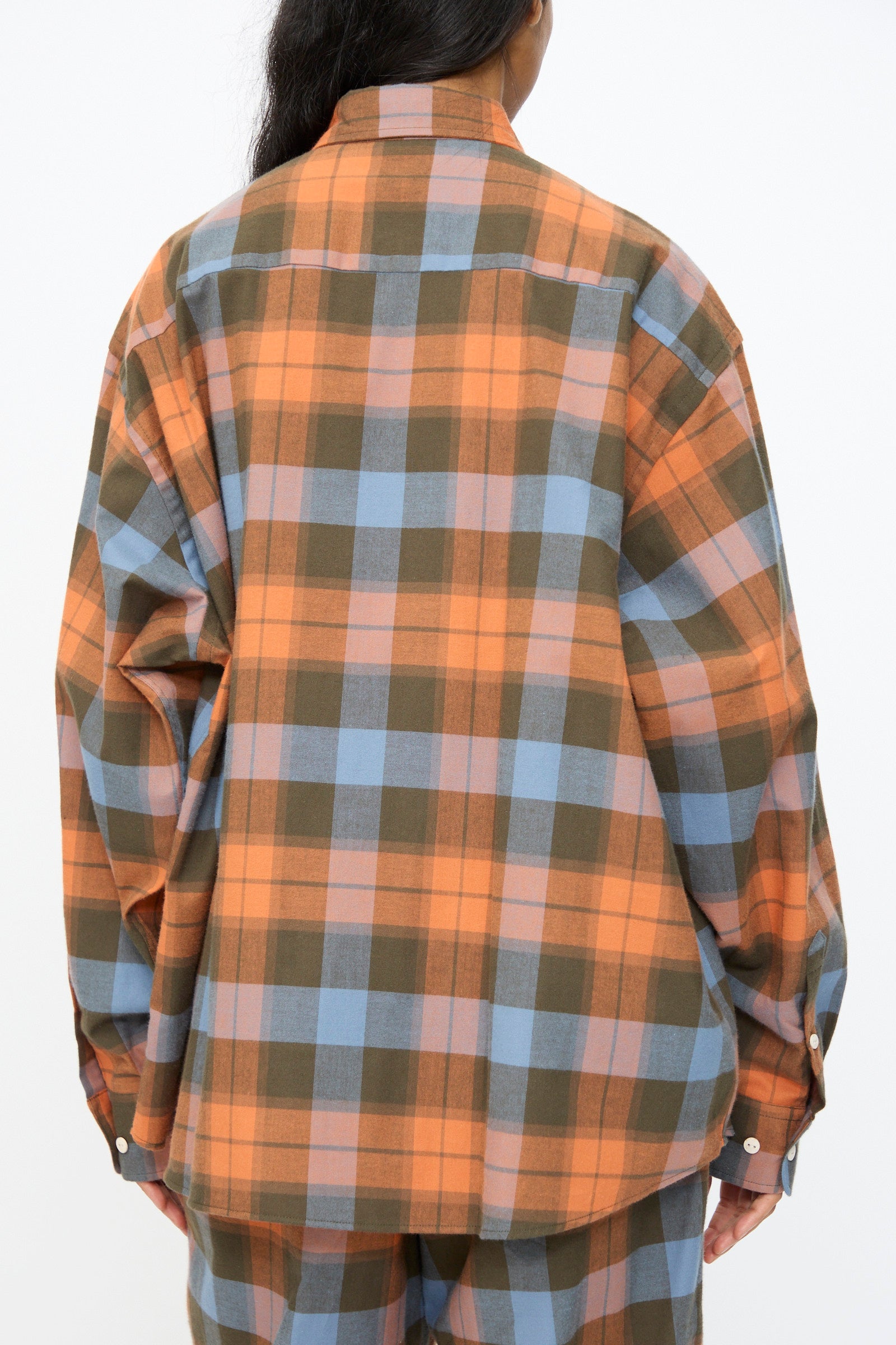 A person is photographed from the back against a plain background, wearing the Baserange Claude Shirt in Frame Plaid—an oversized button-up featuring orange, blue, and green tones.