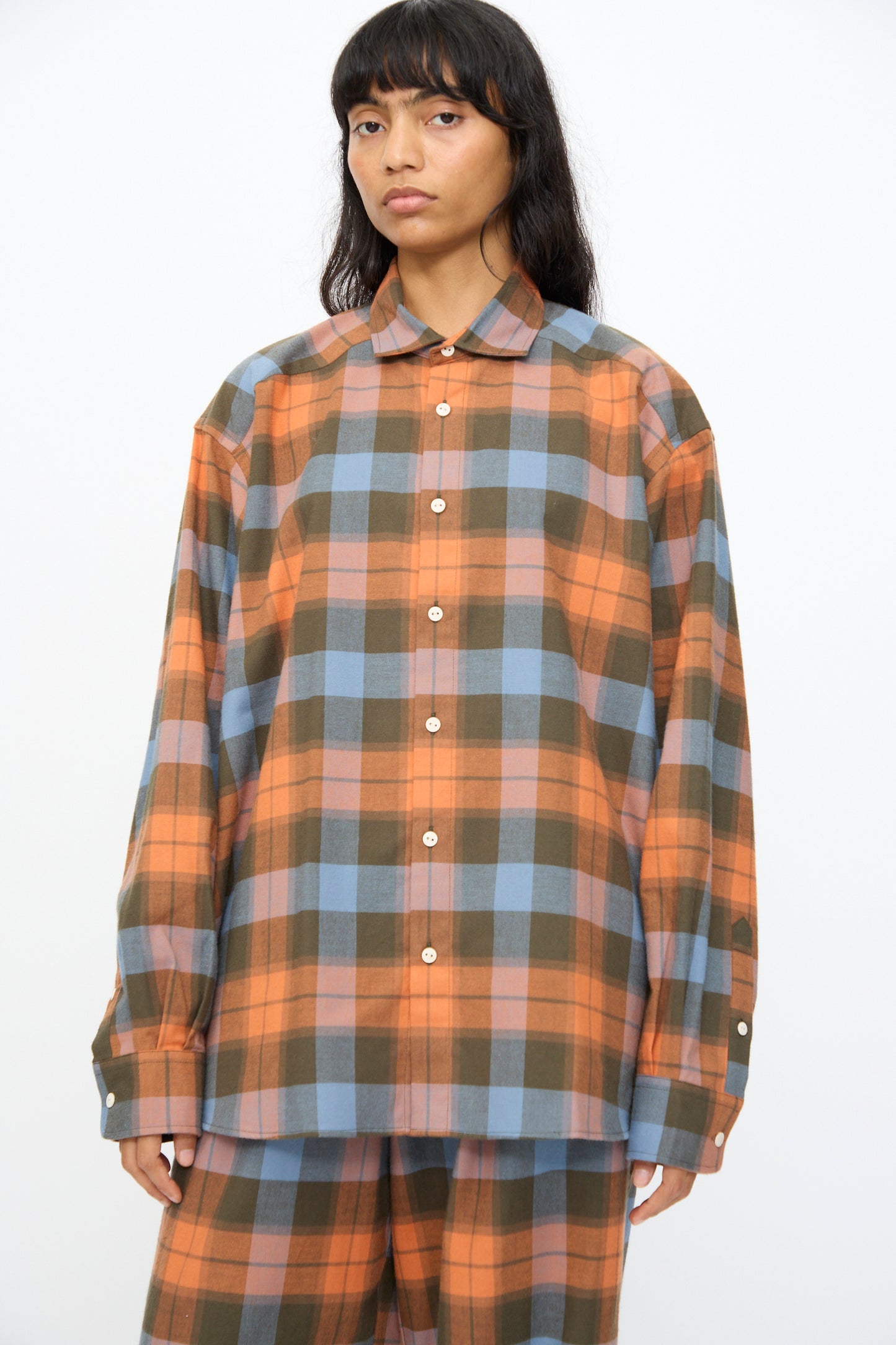 A person wears the Baserange Claude Shirt in Frame Plaid—an oversized button-up with orange, brown, and blue plaid—paired with matching pants, standing against a plain white background.
