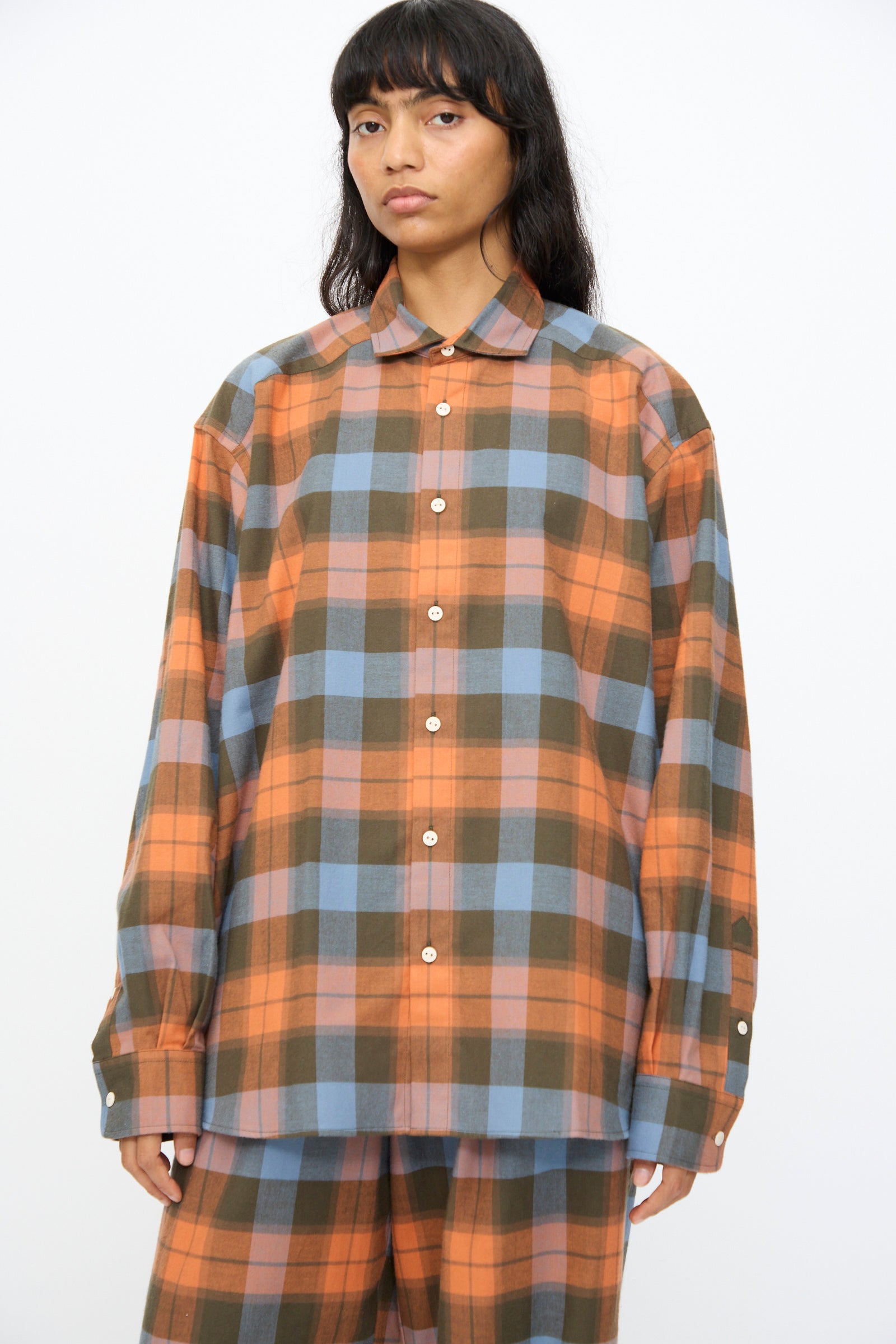 A person wears the Baserange Claude Shirt in Frame Plaid—an oversized button-up with orange, brown, and blue plaid—paired with matching pants, standing against a plain white background.