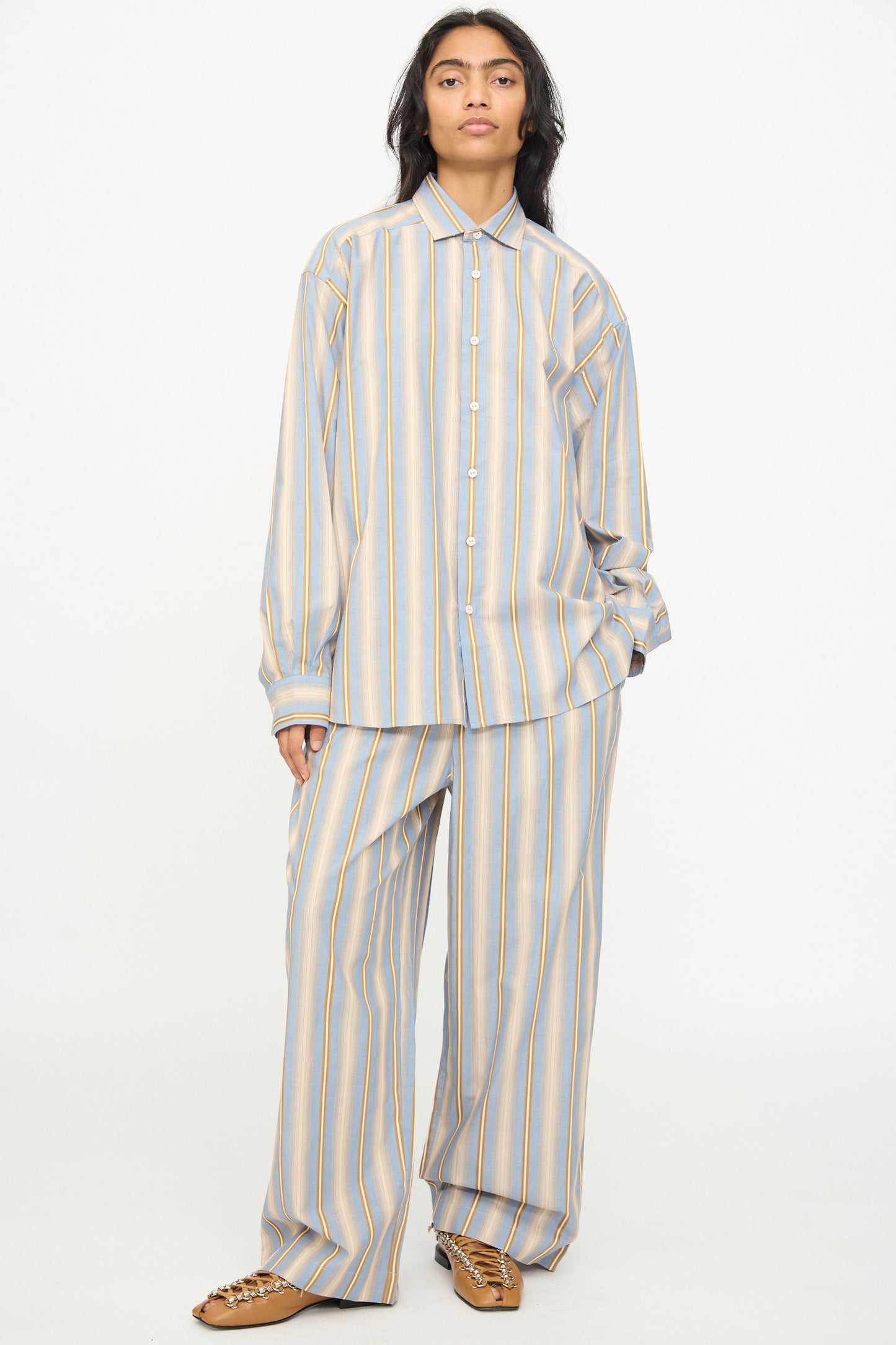 Wearing the Baserange Claude Shirt in Grid Stripe—a relaxed fit, button-up style—paired with wide-leg pants featuring vertical blue, yellow, and white stripes, standing against a plain white background.
