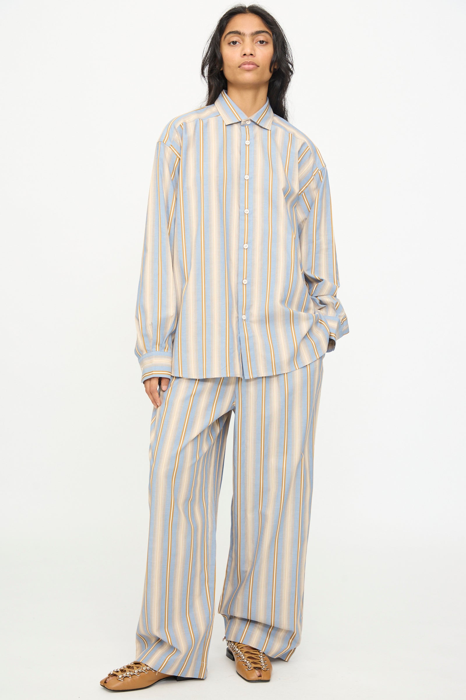 Wearing the Baserange Claude Shirt in Grid Stripe—a relaxed fit, button-up style—paired with wide-leg pants featuring vertical blue, yellow, and white stripes, standing against a plain white background.