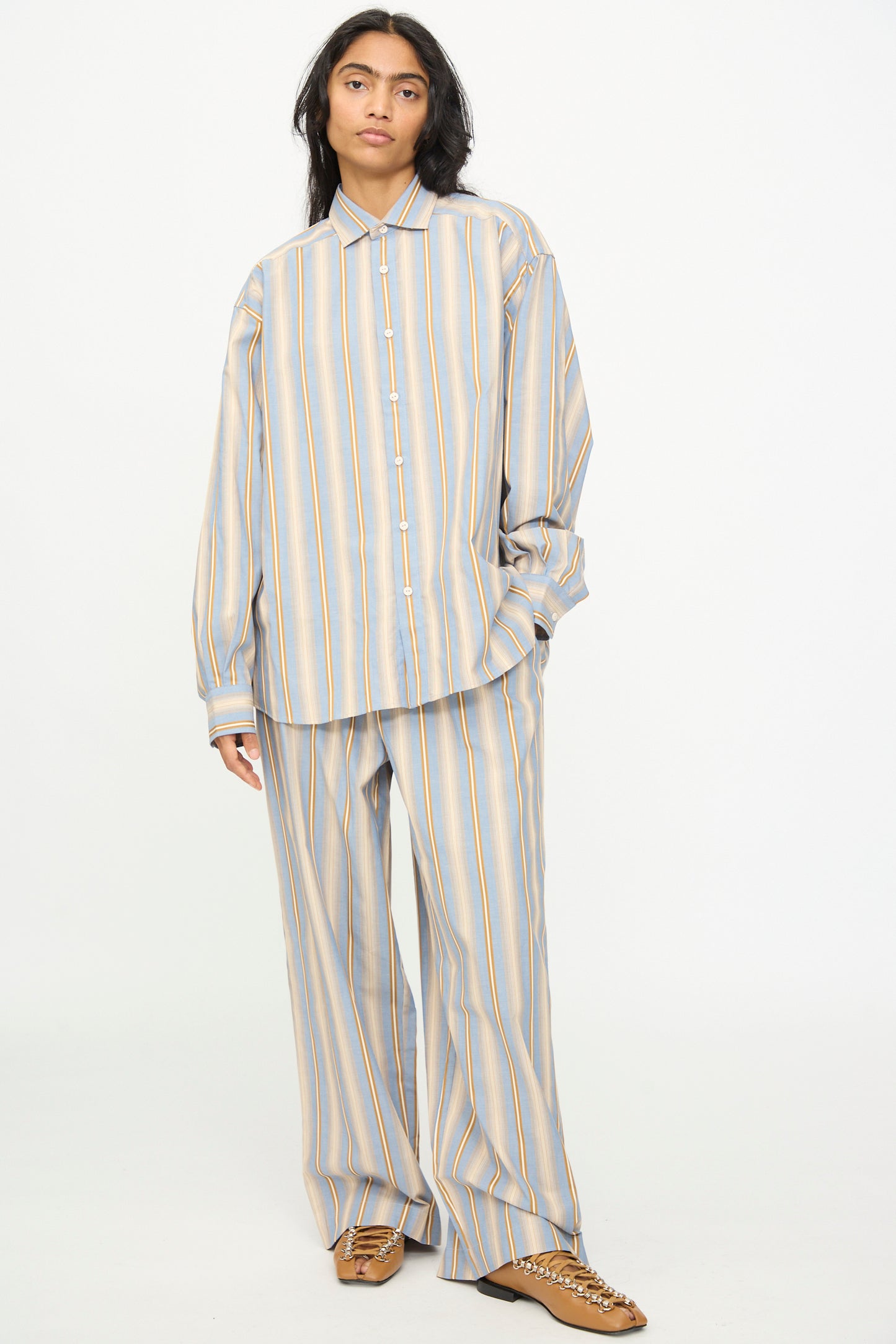 A person wearing the Baserange Claude Shirt in Grid Stripe—a loose-fitting cotton top with matching wide-leg pants featuring muted blue, yellow, and gray stripes—paired with brown shoes, stands against a plain white background.