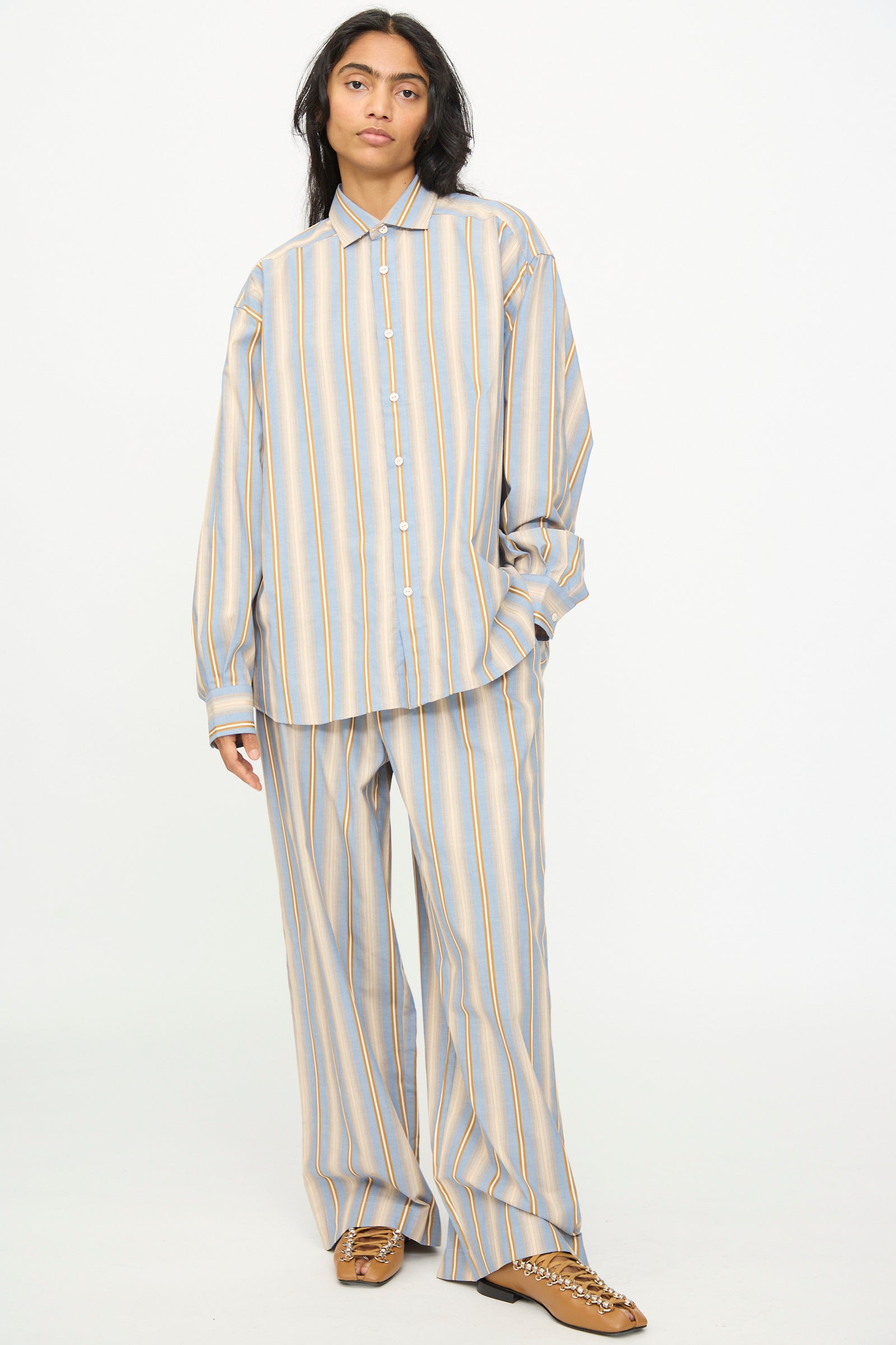 A person wearing the Baserange Claude Shirt in Grid Stripe—a loose-fitting cotton top with matching wide-leg pants featuring muted blue, yellow, and gray stripes—paired with brown shoes, stands against a plain white background.