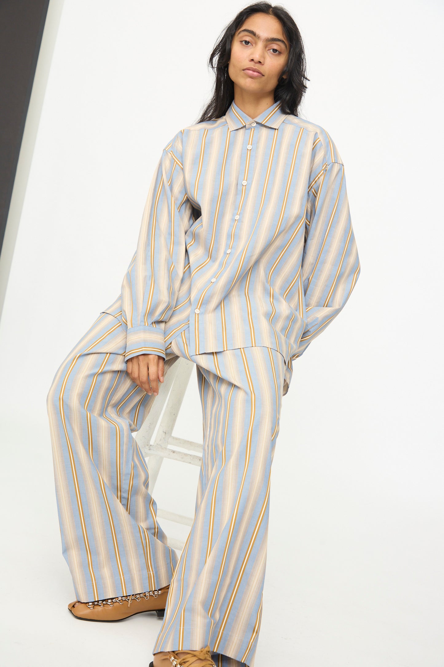 A person sits on a white stool against a plain white background, wearing the Baserange Claude Shirt in Grid Stripe—a relaxed fit, light blue striped cotton shirt with yellow and white vertical stripes—paired with brown shoes.
