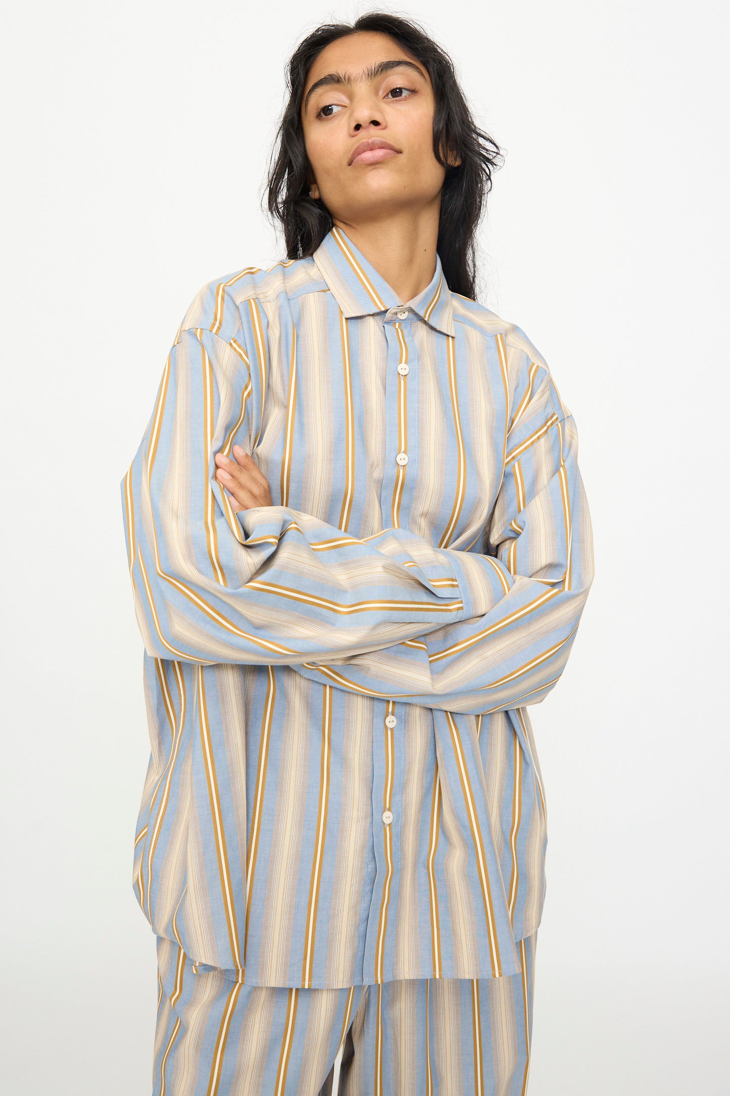 Wearing the Baserange Claude Shirt in Grid Stripe—a loose, relaxed-fit top with matching pants featuring blue, yellow, and white vertical stripes—the person stands against a plain background with arms crossed.