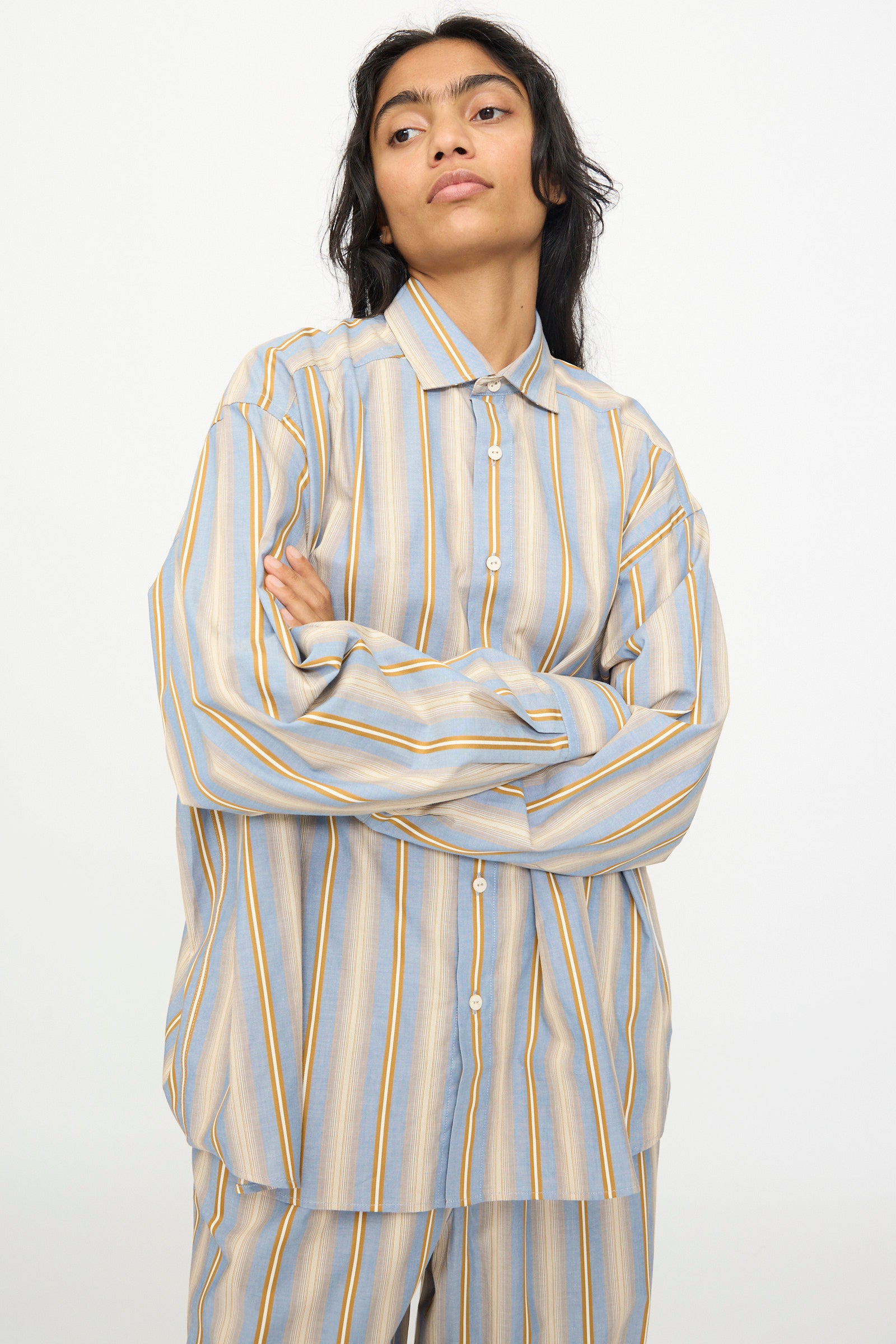 Wearing the Baserange Claude Shirt in Grid Stripe—a loose, relaxed-fit top with matching pants featuring blue, yellow, and white vertical stripes—the person stands against a plain background with arms crossed.