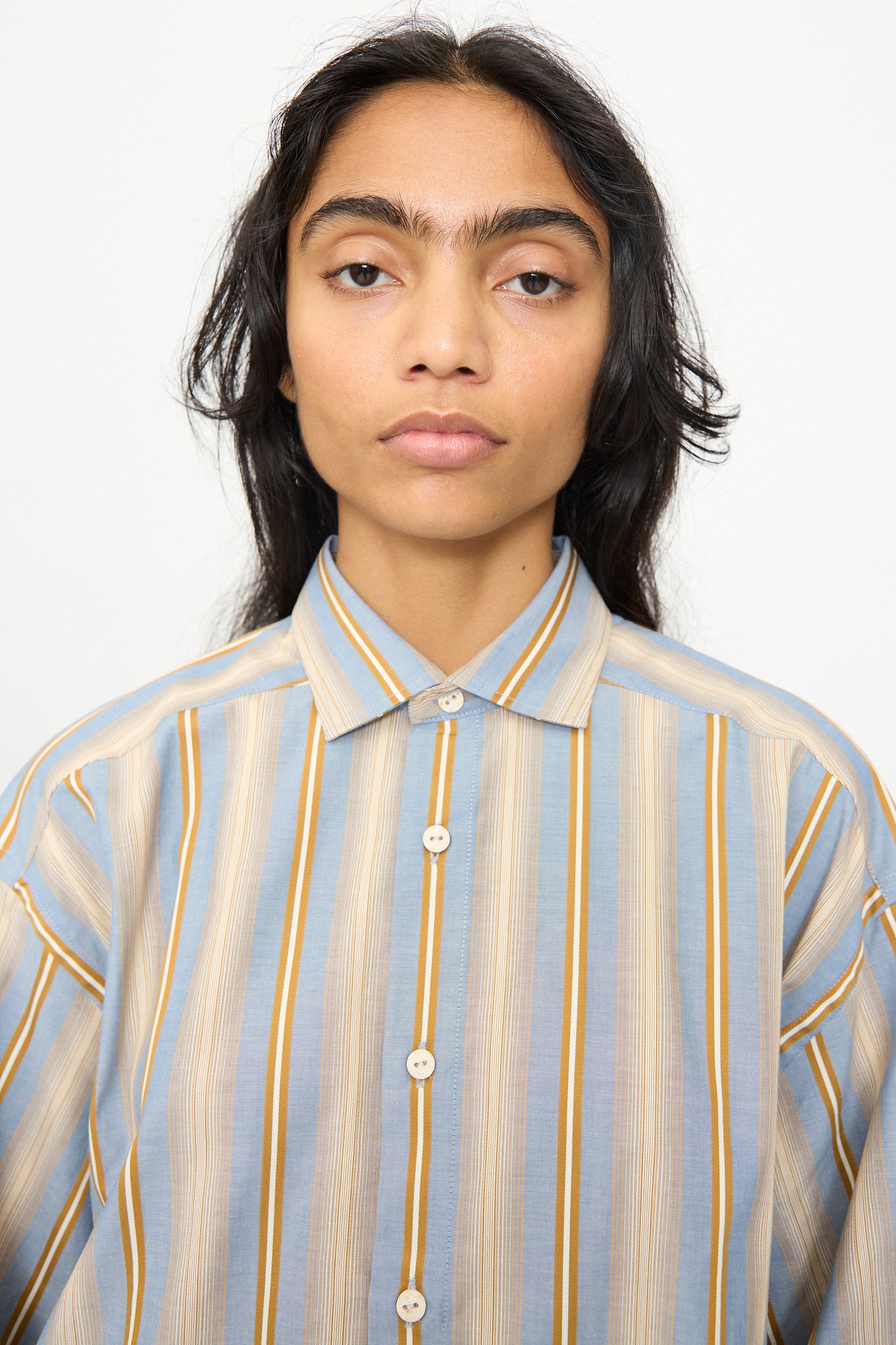 A person with long dark hair and a unibrow faces the camera against a plain white background, wearing the Baserange Claude Shirt in Grid Stripe—a relaxed fit, striped cotton shirt featuring vertical blue, tan, and beige stripes.
