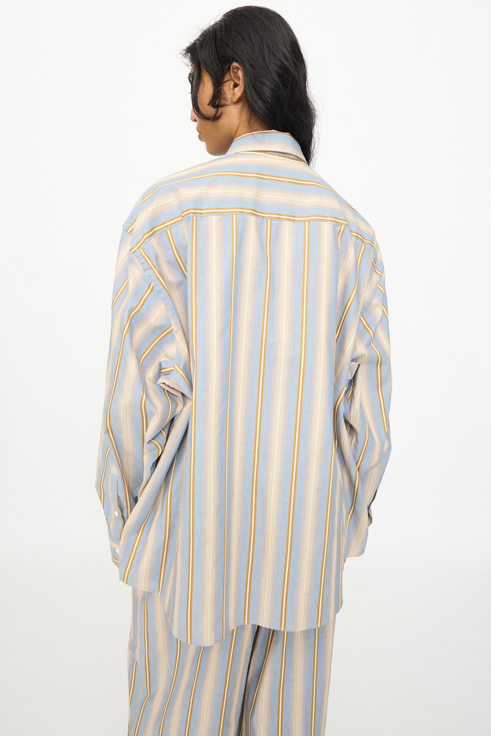 A person with long dark hair faces away, wearing the Baserange Claude Shirt in Grid Stripe—an oversized blue and yellow striped cotton shirt—paired with matching pants against a plain white background.