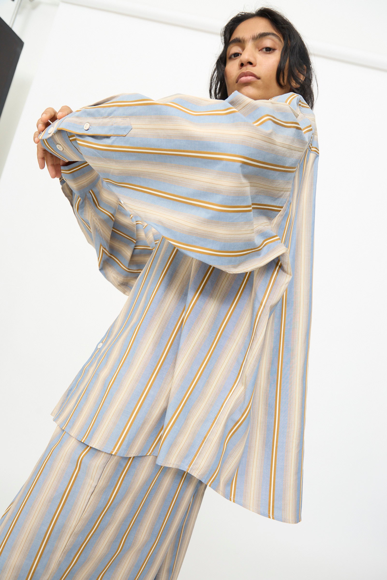 A person wearing the Baserange Claude Shirt in Grid Stripe is photographed from a low angle, holding out one sleeve.