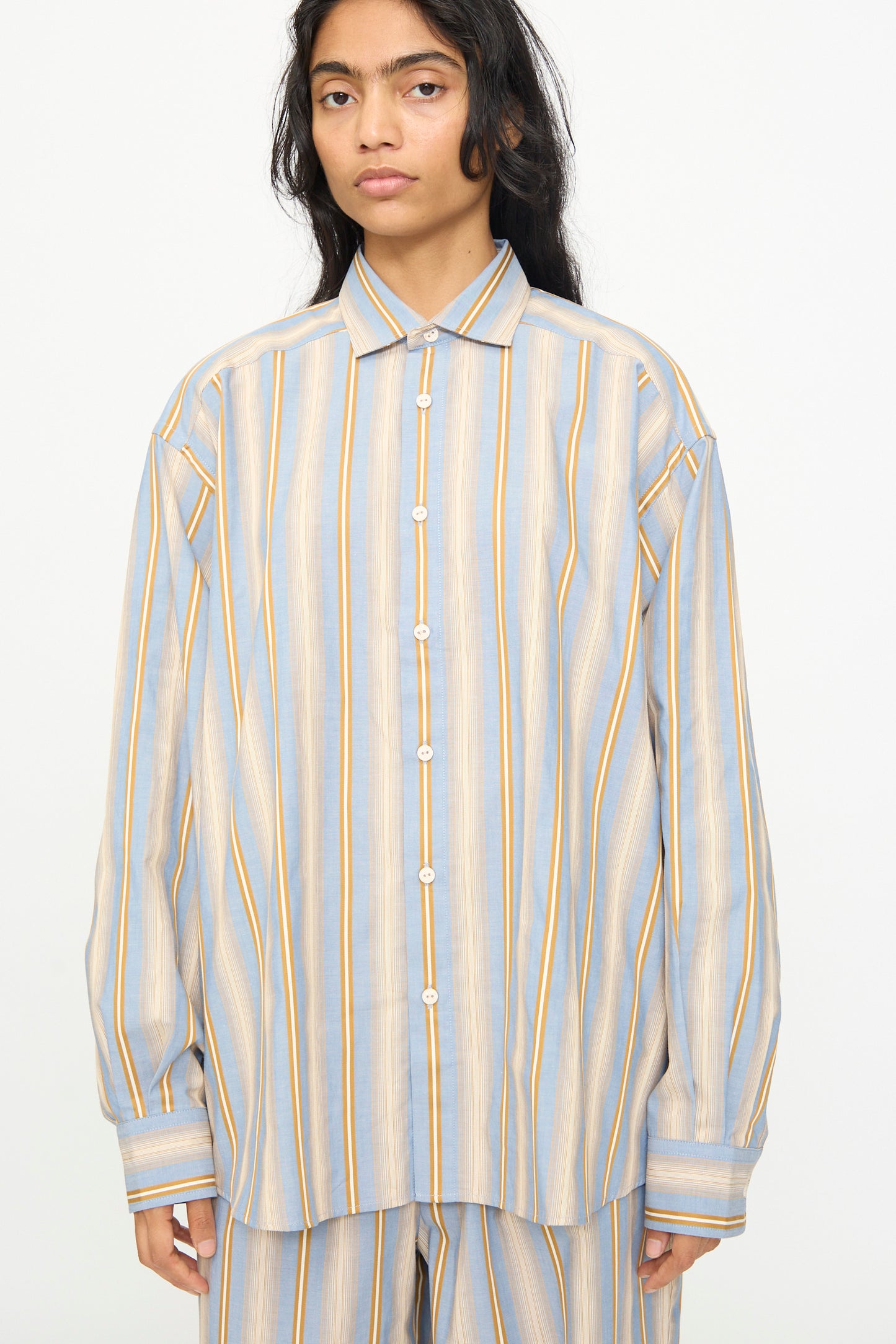 A person wearing the Baserange Claude Shirt in Grid Stripe—an oversized, relaxed fit cotton shirt featuring long sleeves and vertical blue, beige, and yellow stripes—stands in front of a plain white background.