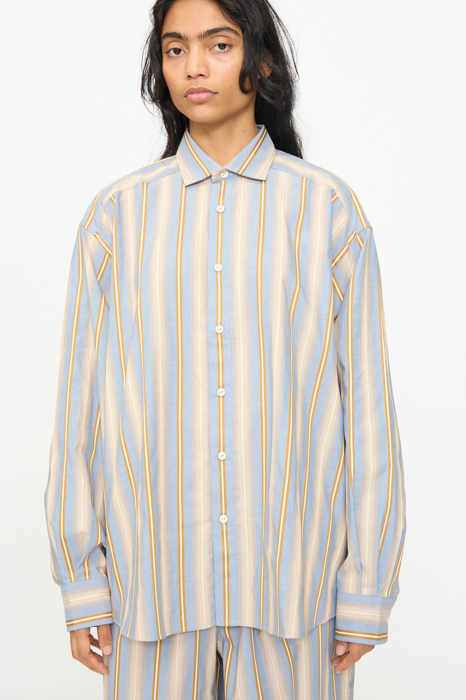 A person wearing the Baserange Claude Shirt in Grid Stripe—an oversized, relaxed fit cotton shirt featuring long sleeves and vertical blue, beige, and yellow stripes—stands in front of a plain white background.