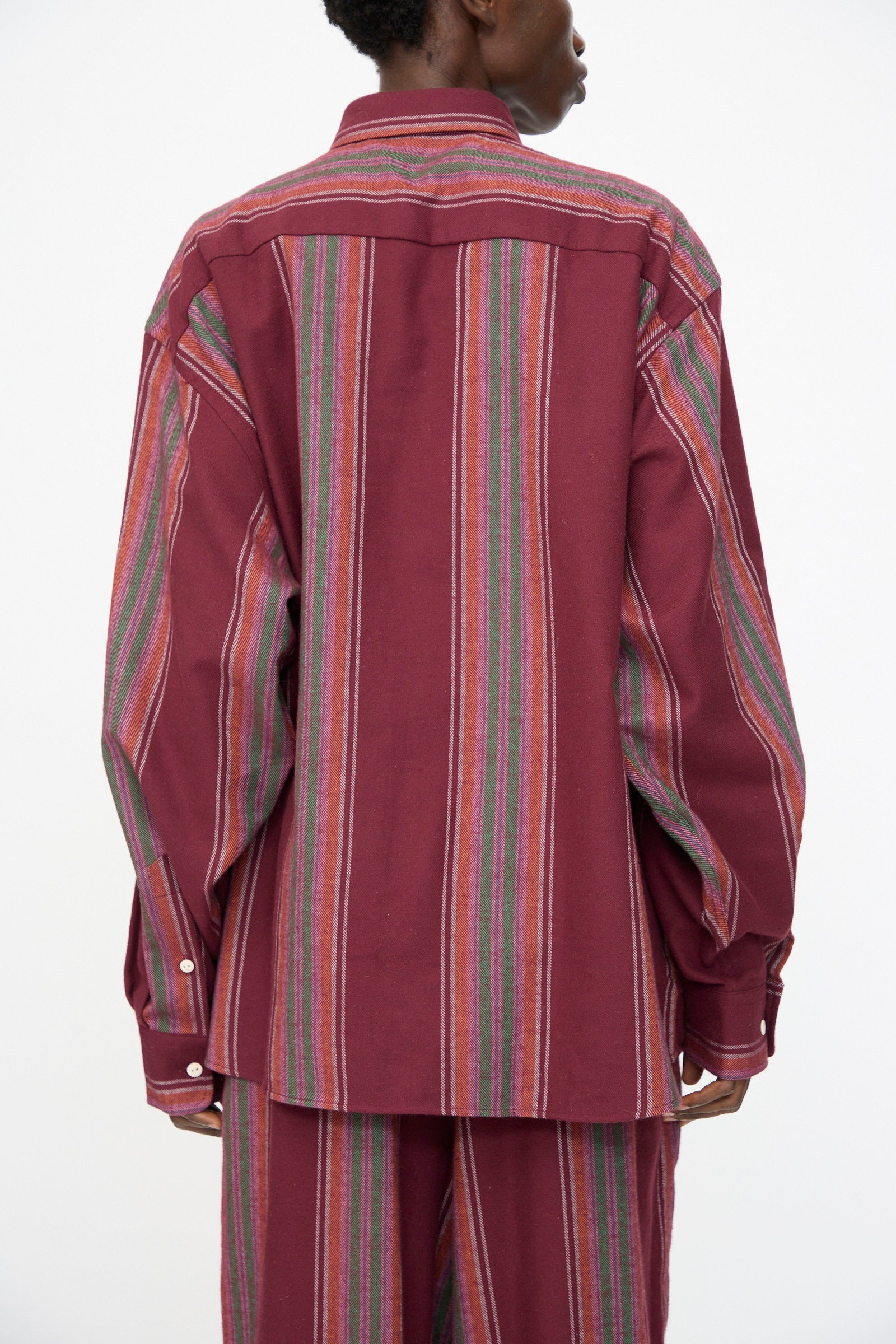 A person is shown from the back wearing the Baserange Claude Shirt in Maroon Stripe, a loose-fitting, long-sleeve organic cotton shirt with multicolored vertical accents; matching pants are partially visible.