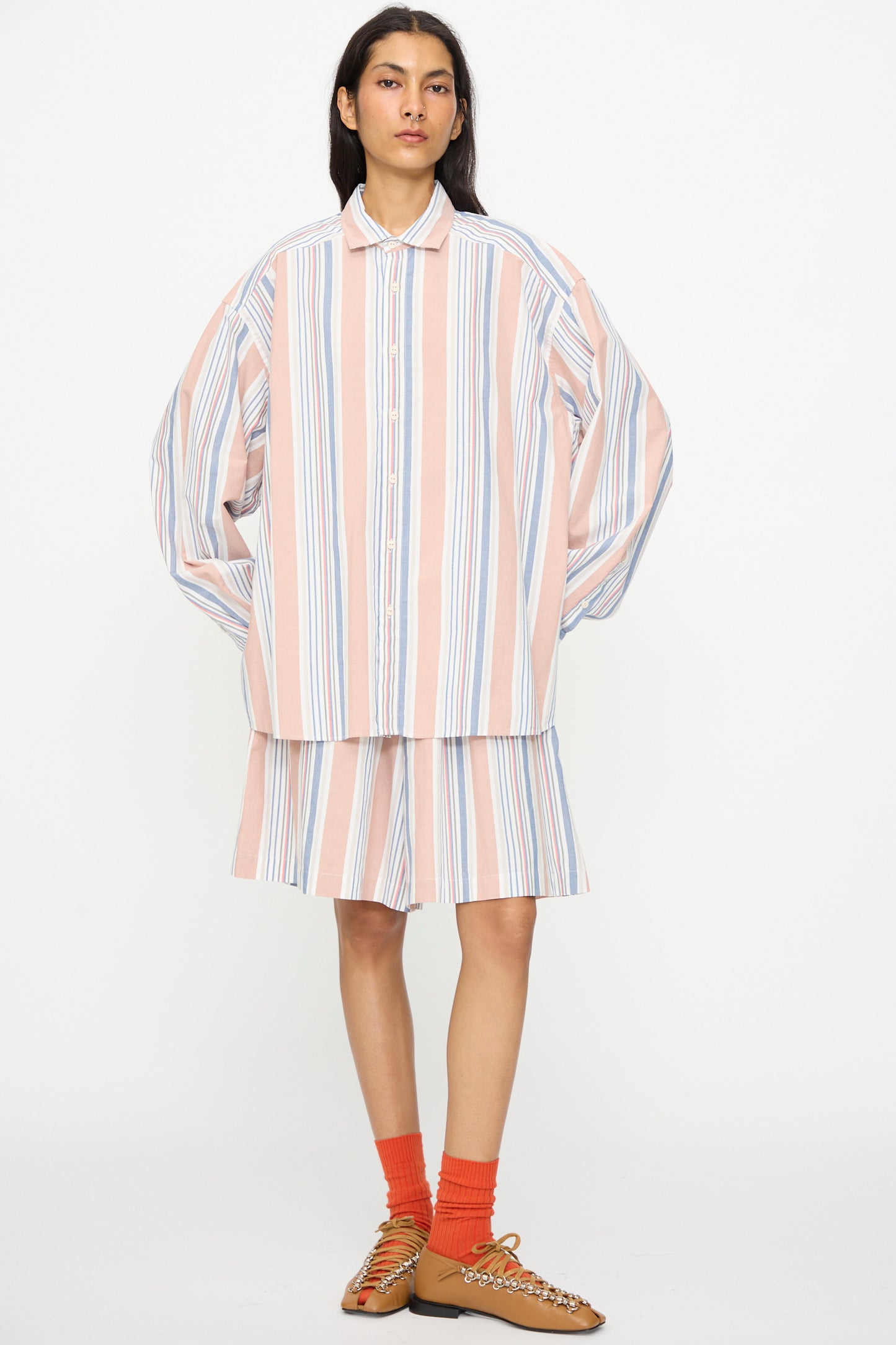 A person stands against a white background wearing Baserange's Claude Shorts in Martinique Brick—a loose, striped, cotton short with an elastic waistband—paired with a matching shirt, orange socks, and brown shoes.