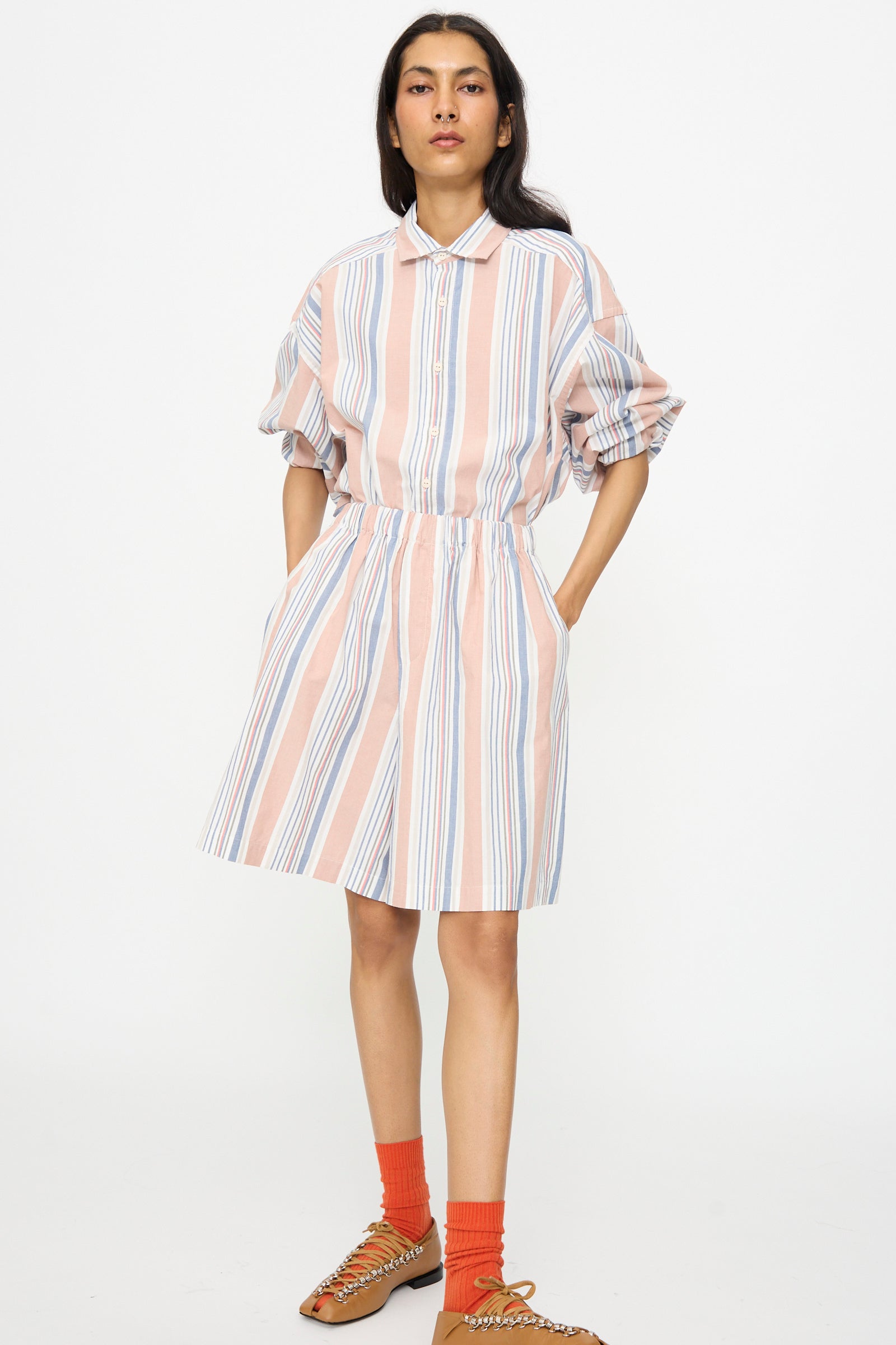 A person wears a collared, short-sleeve dress with vertical pink, white, and blue stripes over Baserange Claude Shorts in Martinique Brick, paired with orange socks and tan woven sandals, styled against a plain white background.