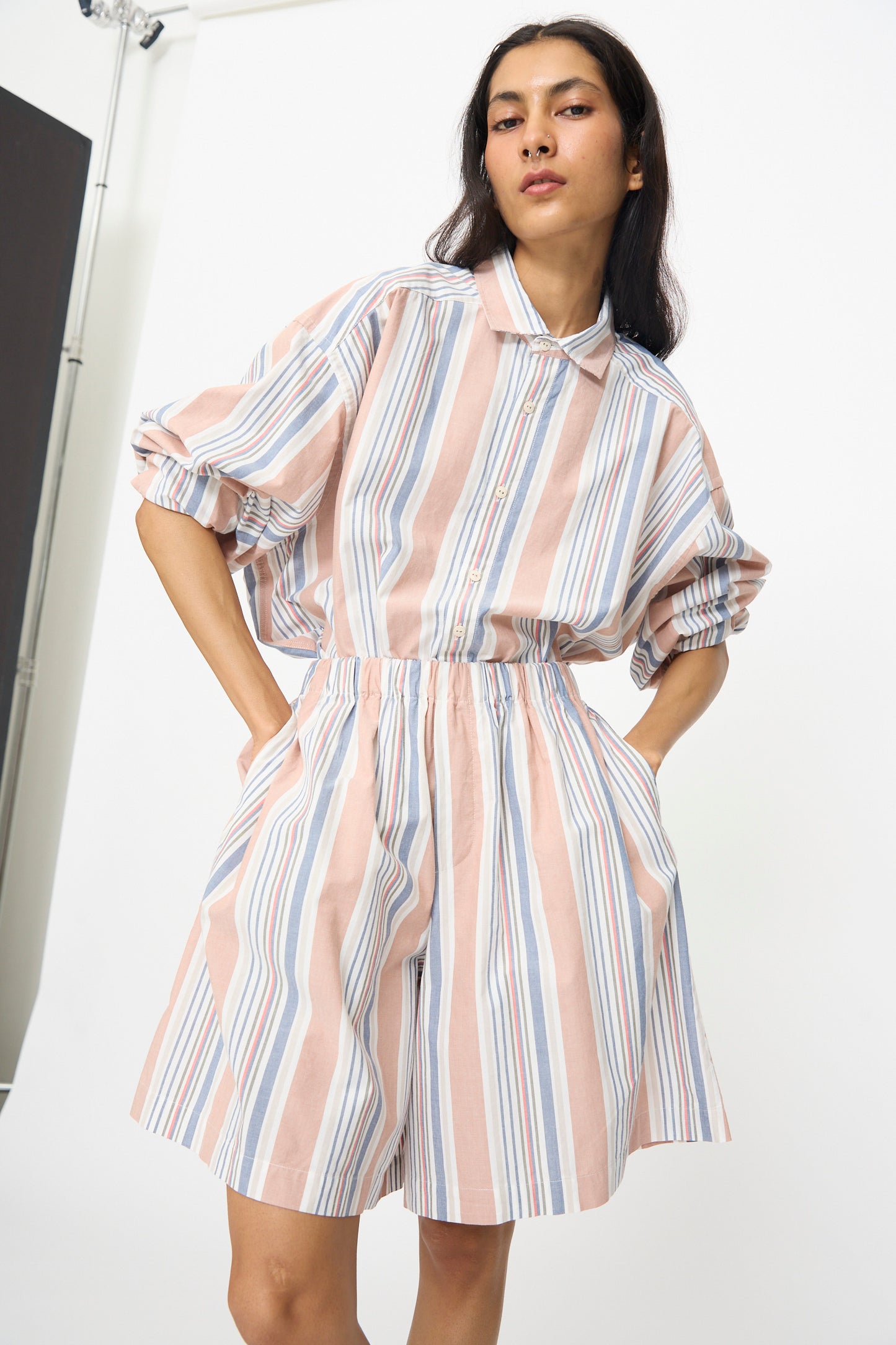 A woman stands with her hands in her pockets against a white background, wearing Baserange's Claude Shorts in Martinique Brick and a loose-fitting, pastel striped button-up shirt.
