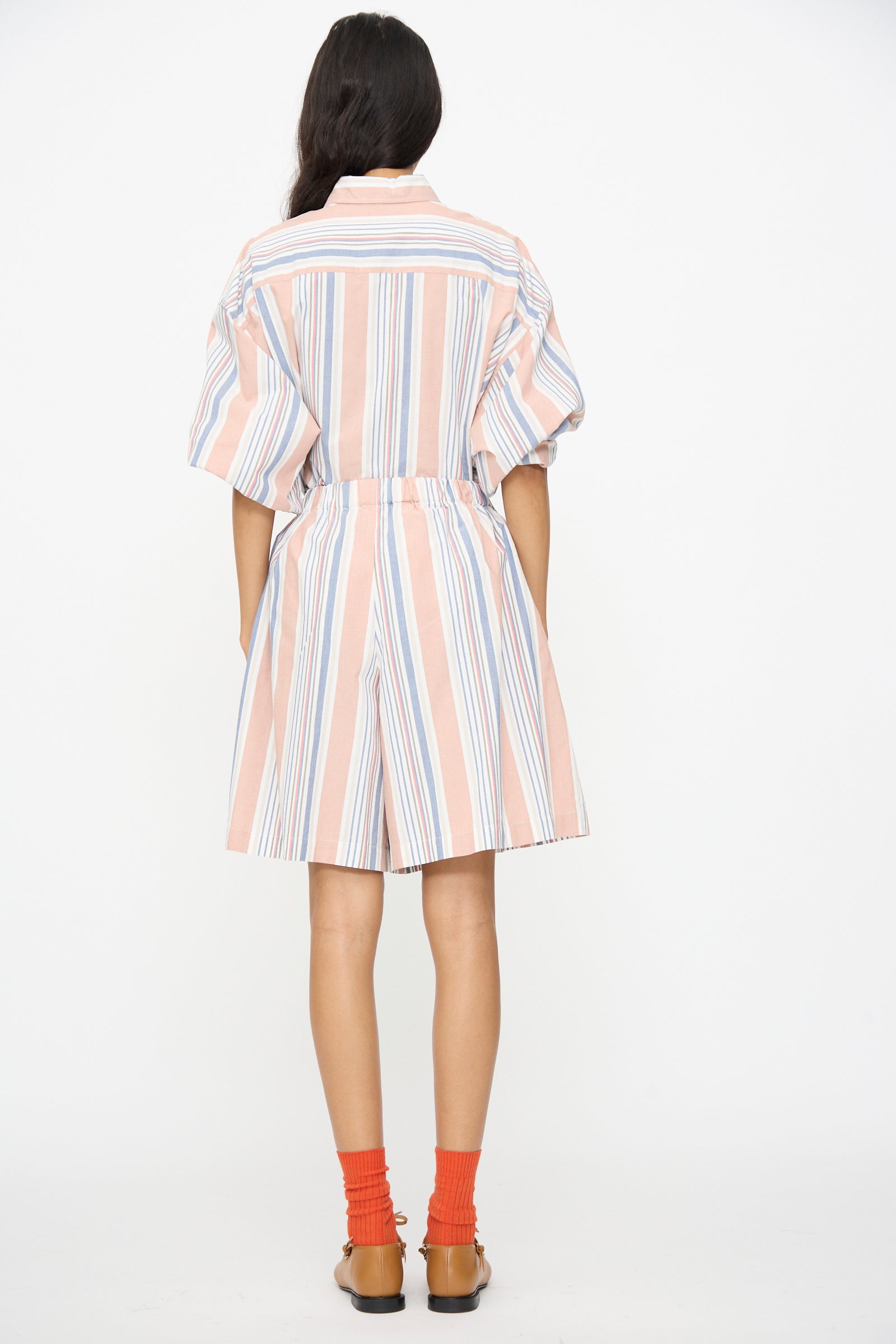 Person stands facing away, wearing a short-sleeved, knee-length dress with vertical pastel stripes, orange socks, brown sandals, and Baserange Claude Shorts in Martinique Brick peeking out from beneath the dress.