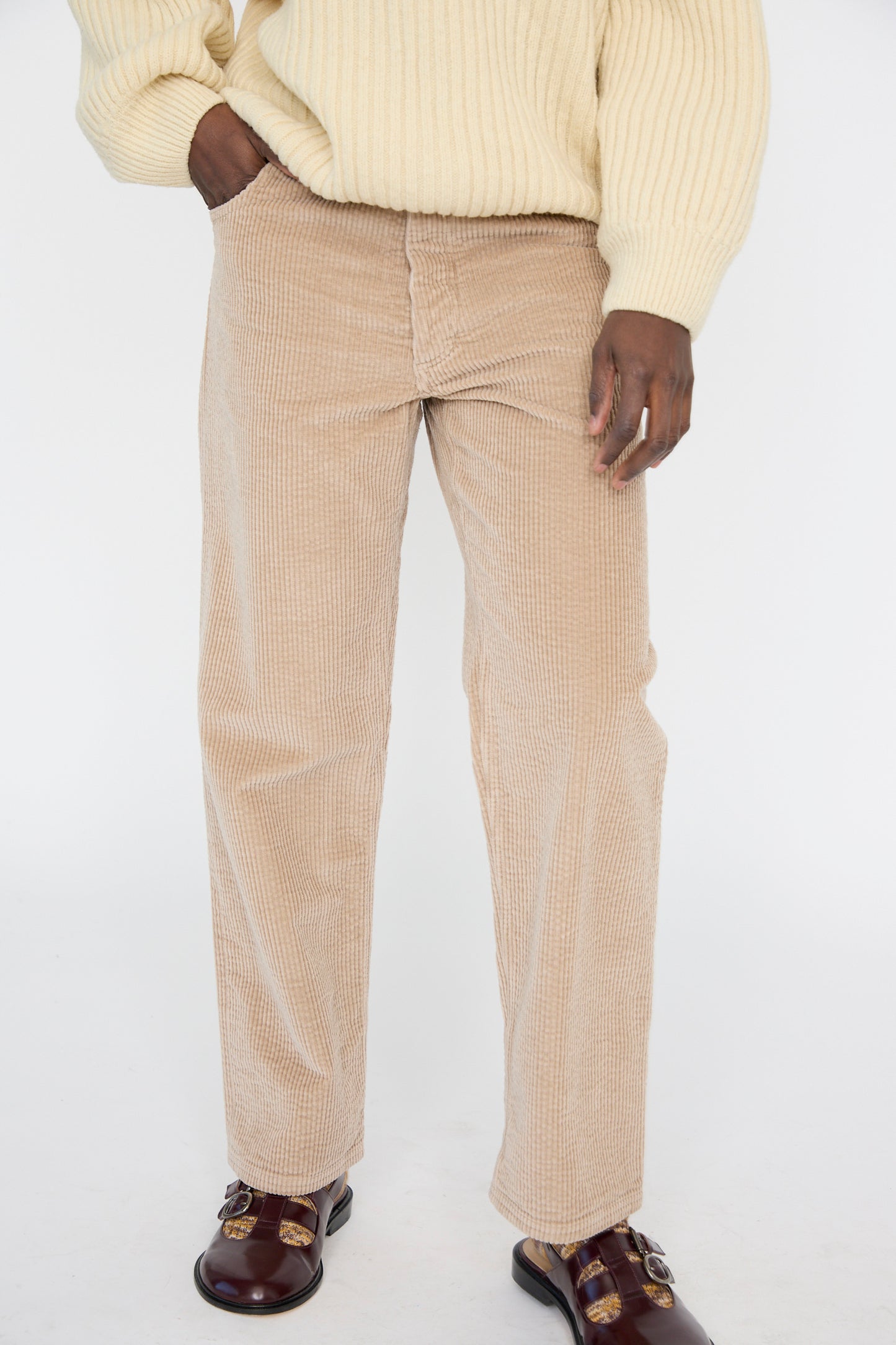 A model wearing a cream sweater, relaxed fit Corduroy Indre Pant in Sand by Baserange, and brown shoes with buckles seen from below the waist. 