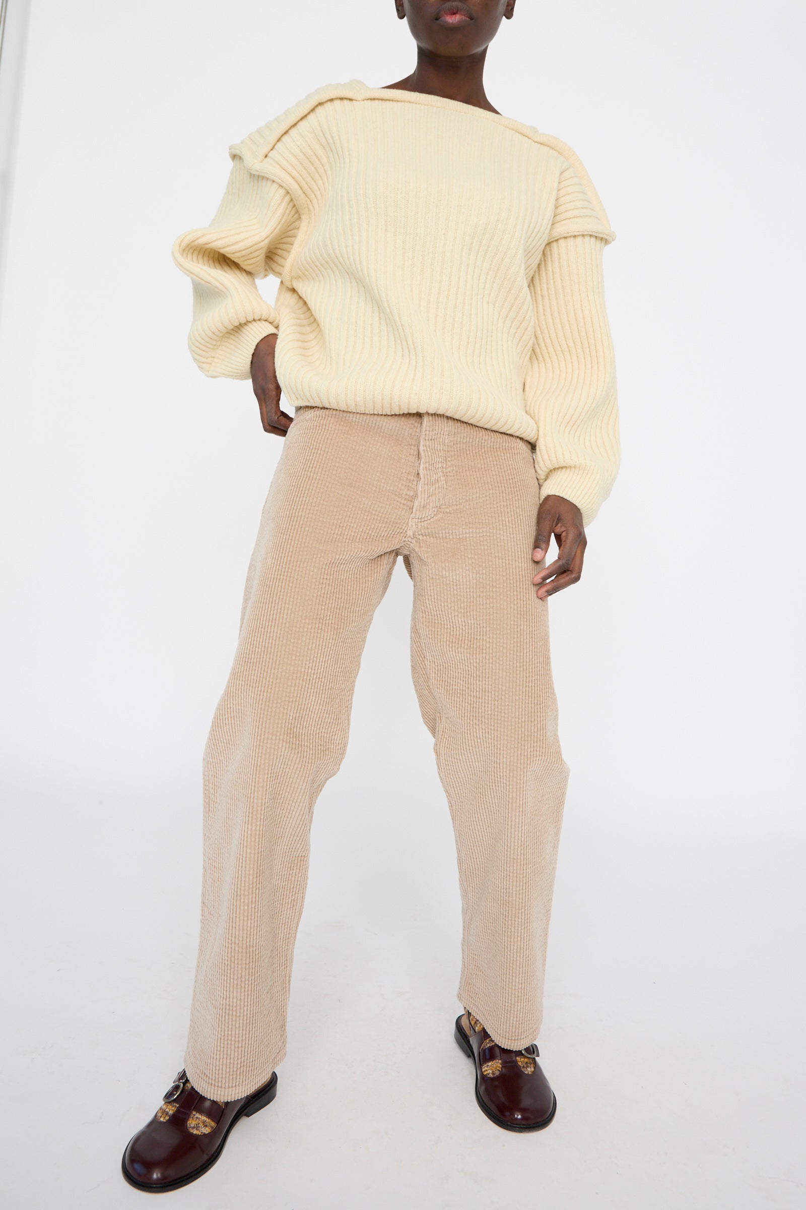 A person wearing a cream oversized sweater, Baserange's relaxed-fit Corduroy Indre Pant in Sand, and brown loafers stands against a plain white background.