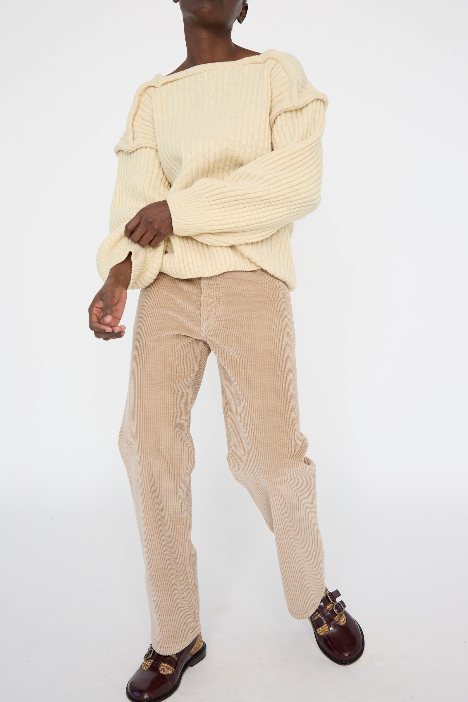 A person wearing a cream knit sweater, Corduroy Indre Pant in Sand by Baserange, and brown loafers against a white background.