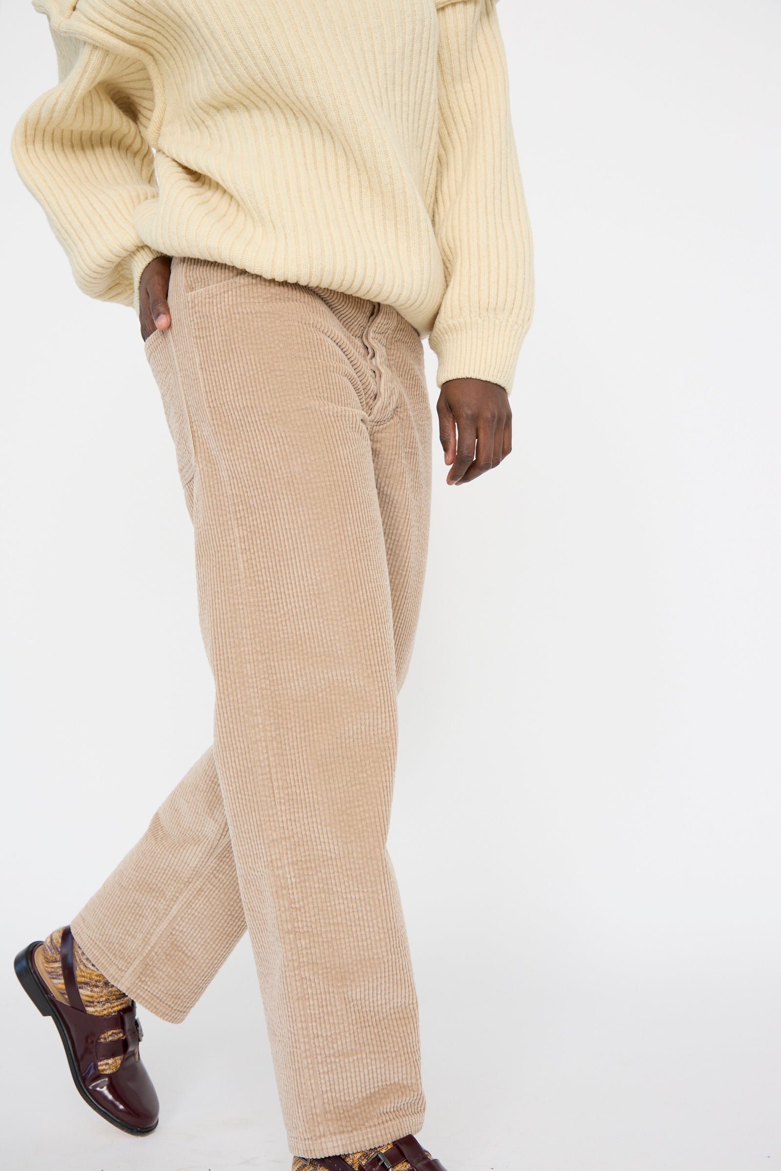 A person wearing a cream sweater and Baserange's Corduroy Indre Pant in Sand with brown shoes walks forward against a plain background.