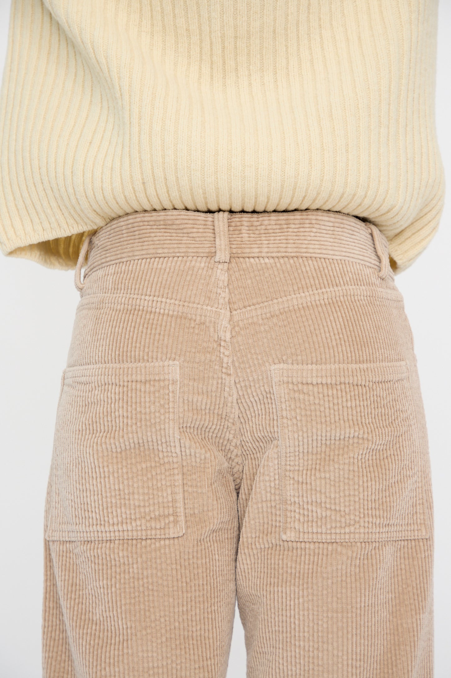 A person is facing away wearing a vertically ribbed beige sweater and Baserange Corduroy Indre Pant in Sand, featuring visible back pockets, closeup on the hips. 