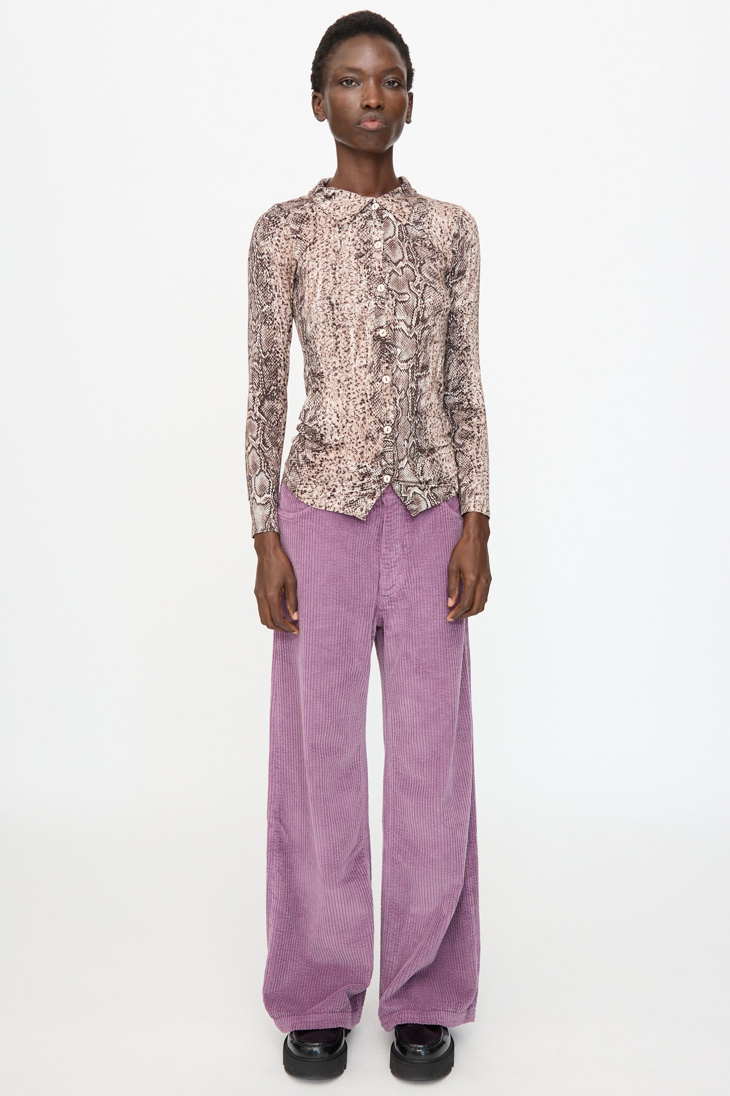 A person stands facing forward against a plain white background, wearing the Baserange Cosme Shirt in Snake, paired with wide-leg purple corduroy pants and black platform shoes.