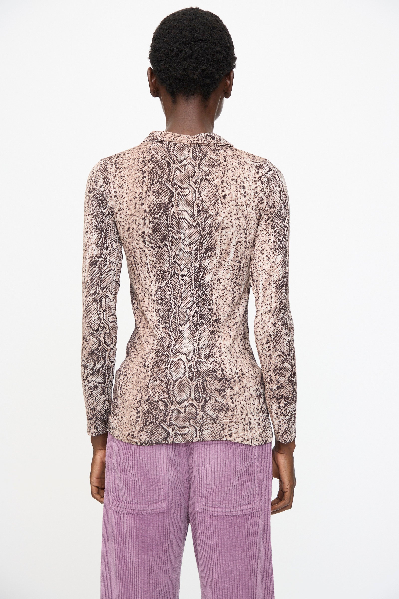 A person faces away, wearing the Baserange Cosme Shirt in Snake, a slim fit brown snakeskin-patterned long sleeve shirt, paired with light purple corduroy pants against a plain white background.