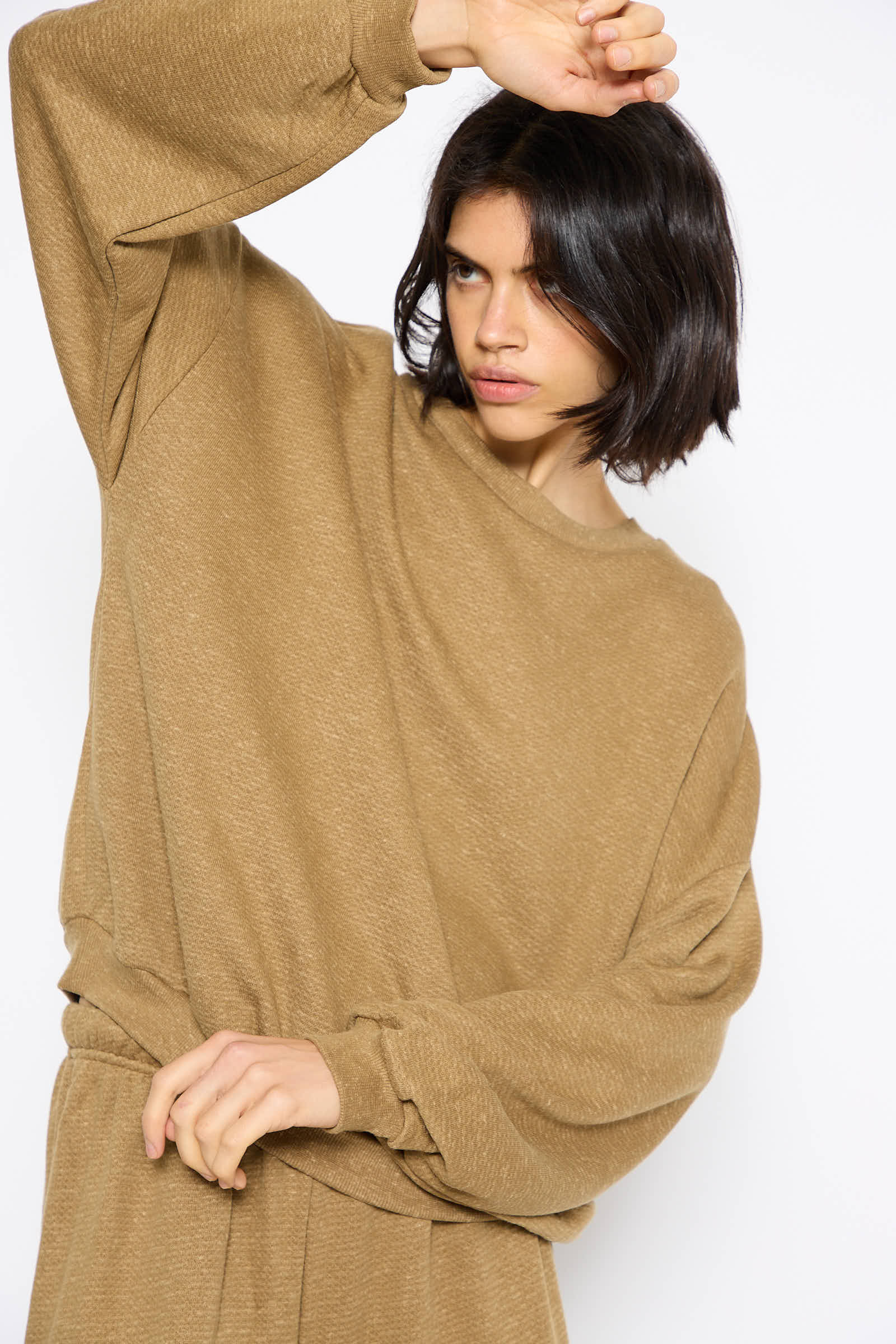 A person with short dark hair wearing a loose-fitting, oversized Baserange Cotton Fleece Rim Sweatshirt in Stome Green made from regenerative cotton is posing with one arm above their head.