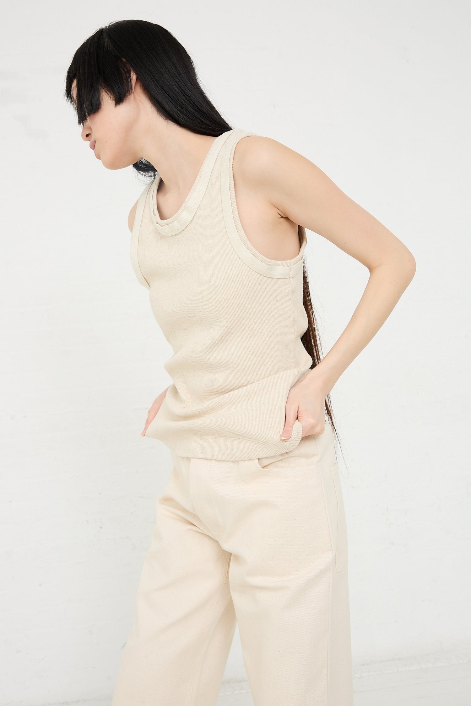 A woman with black hair wearing a beige Baserange Organic Cotton Hemp Rib Supple Tank in Undyed and pants stands in a profile pose against a white background.