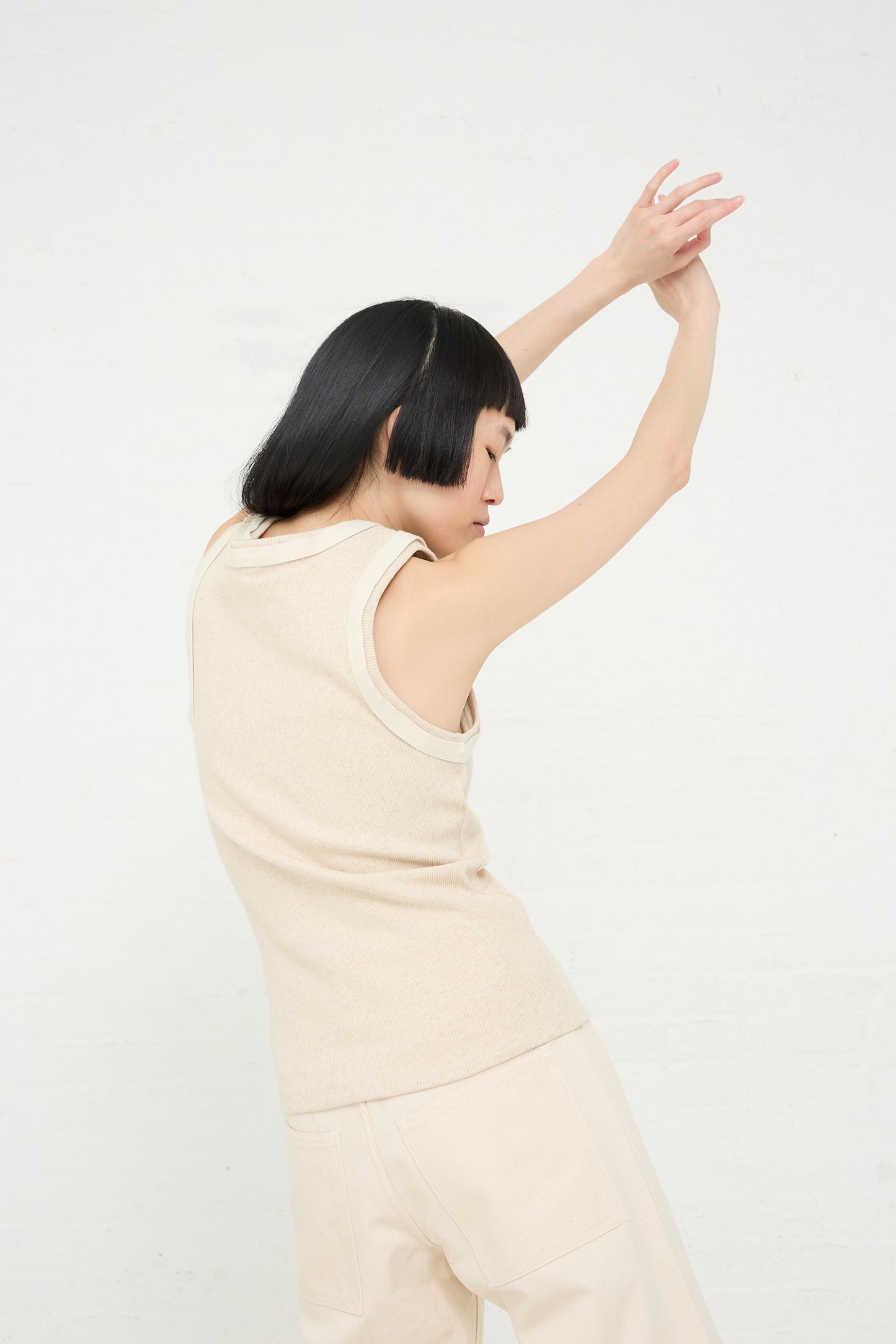 A woman with short dark hair dressed in a Organic Cotton Hemp Rib Supple Tank in Undyed from Baserange performs a dance move against a white background.