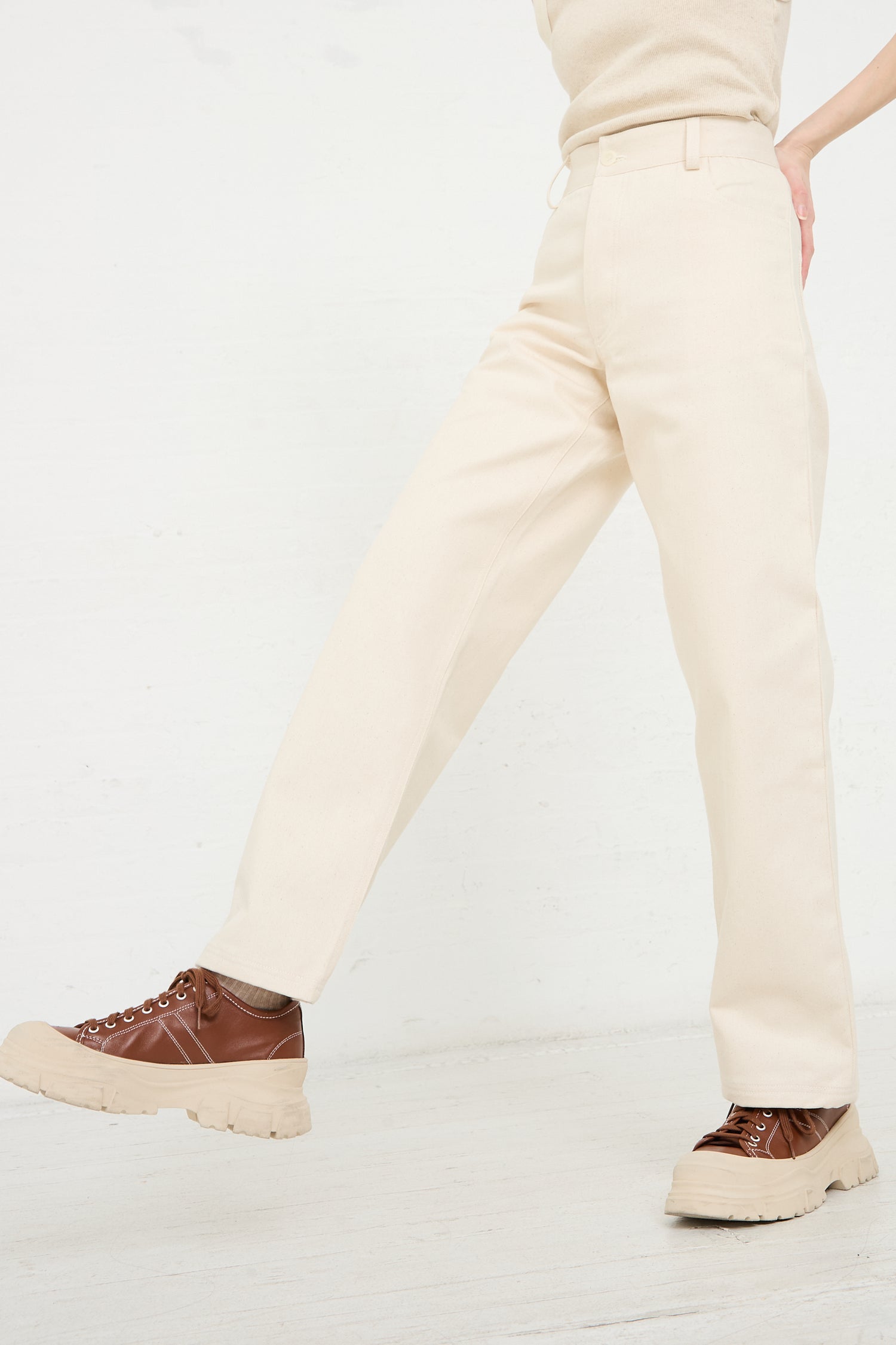 Person wearing Baserange's Organic Cotton Indre Pant in Undyed and brown chunky lace-up shoes walking against a white background.