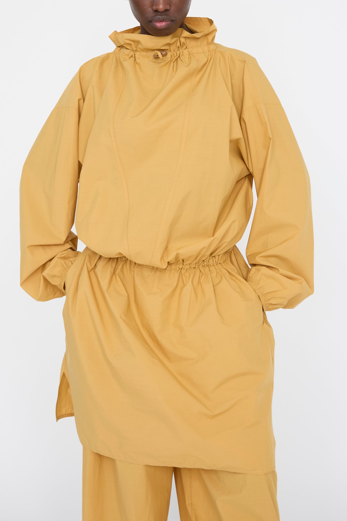 A person is dressed in a Baserange Cotton Poplin Air Dress in Sighs Yellow, featuring an oversized fit with organic cotton, a gathered waist, high collar, and long sleeves, with hands in the pockets. 