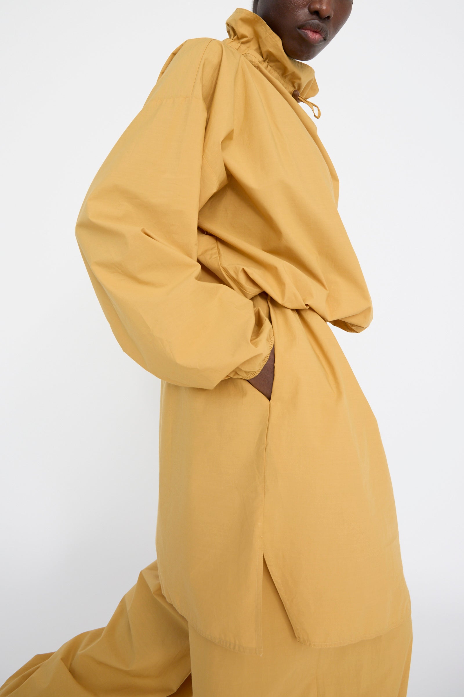 A person is wearing the Baserange Cotton Poplin Air Dress in Sighs Yellow, featuring a mustard yellow midi design with a high collar and loose sleeves. Made from organic cotton, it boasts an oversized fit with a cinched waist and two side slits.