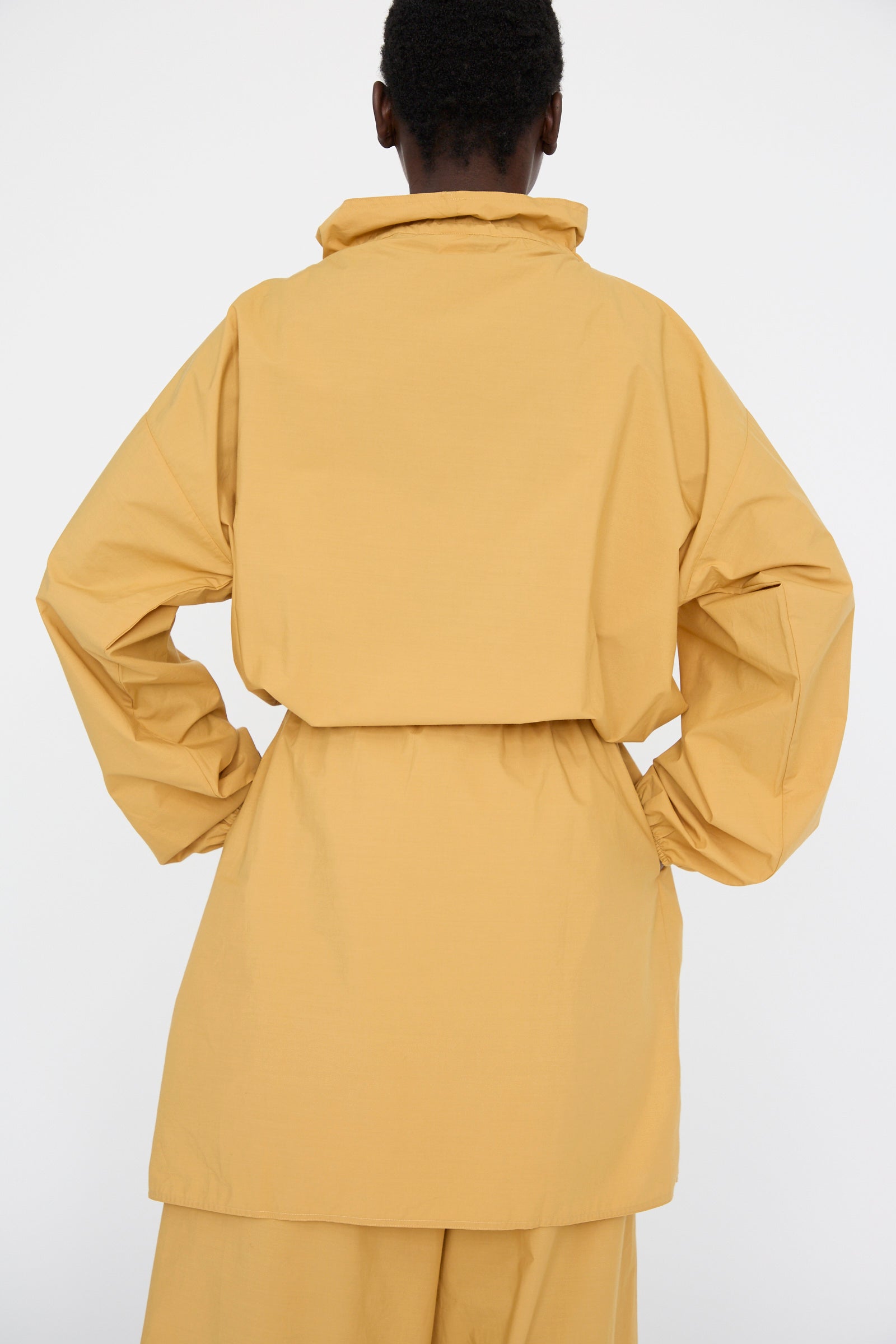 A person wearing a Baserange Cotton Poplin Air Dress in Sighs Yellow stands with hands on hips, turned away from the camera against a white background. 