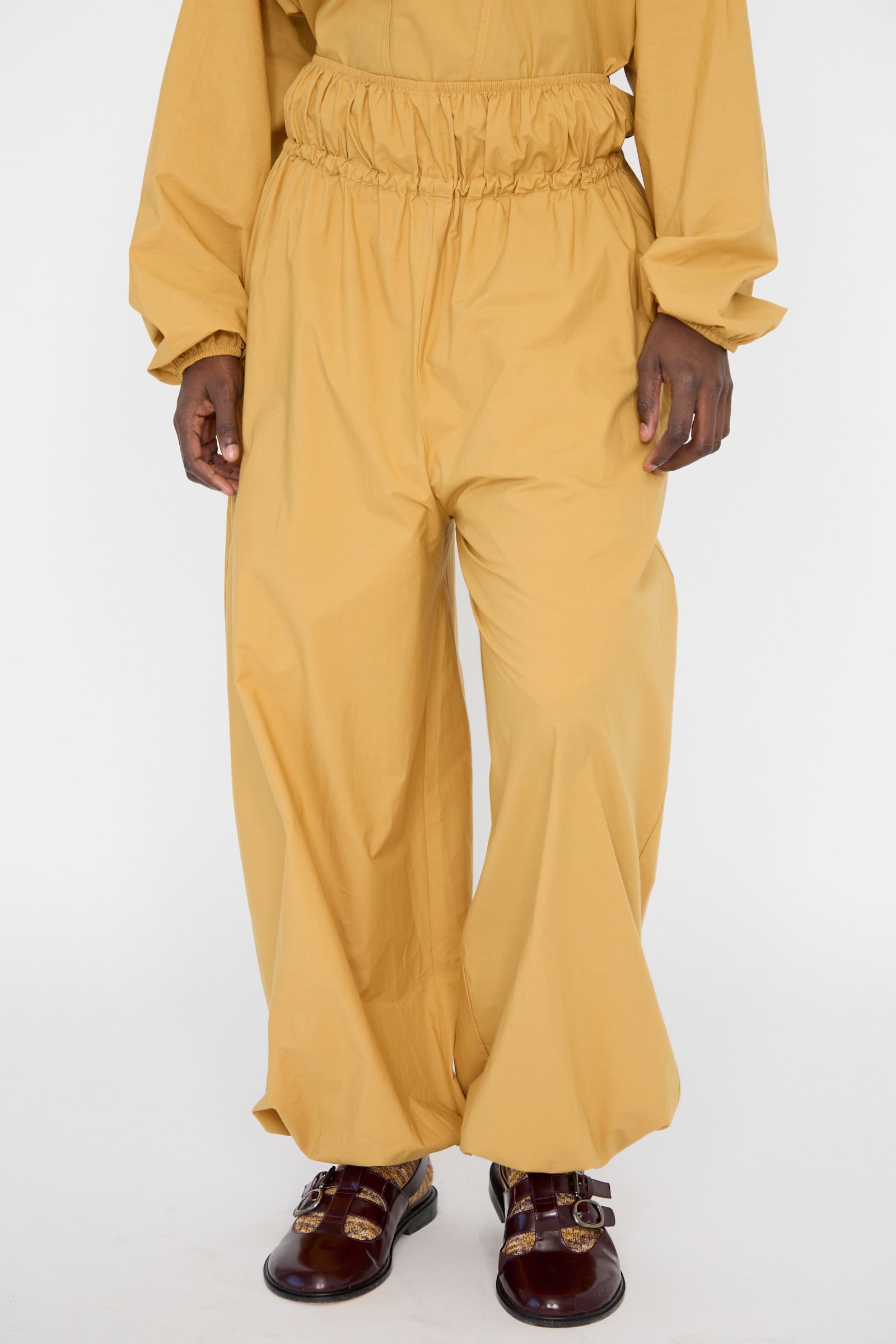 Person wearing the Cotton Poplin Air Pants in Sighs Yellow by Baserange and burgundy shoes against a white backdrop, seen from below the shoulders. 