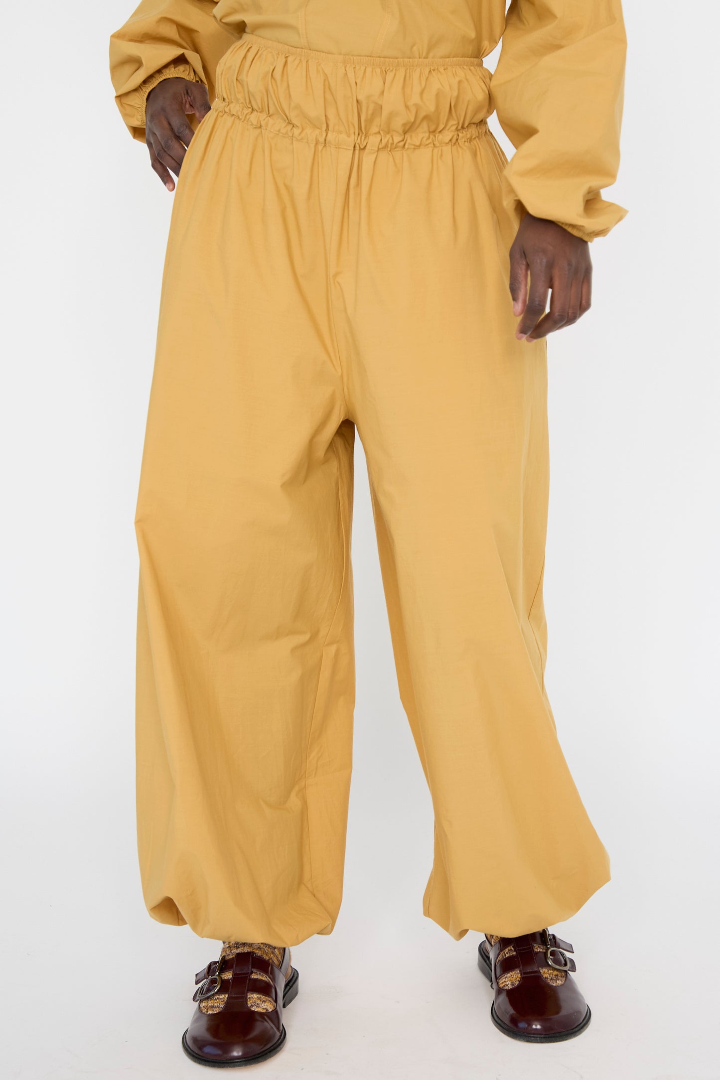 A person wearing Baserange's Cotton Poplin Air Pants in Sighs Yellow, paired with brown buckle shoes, viewed from below the hips against a plain background. 