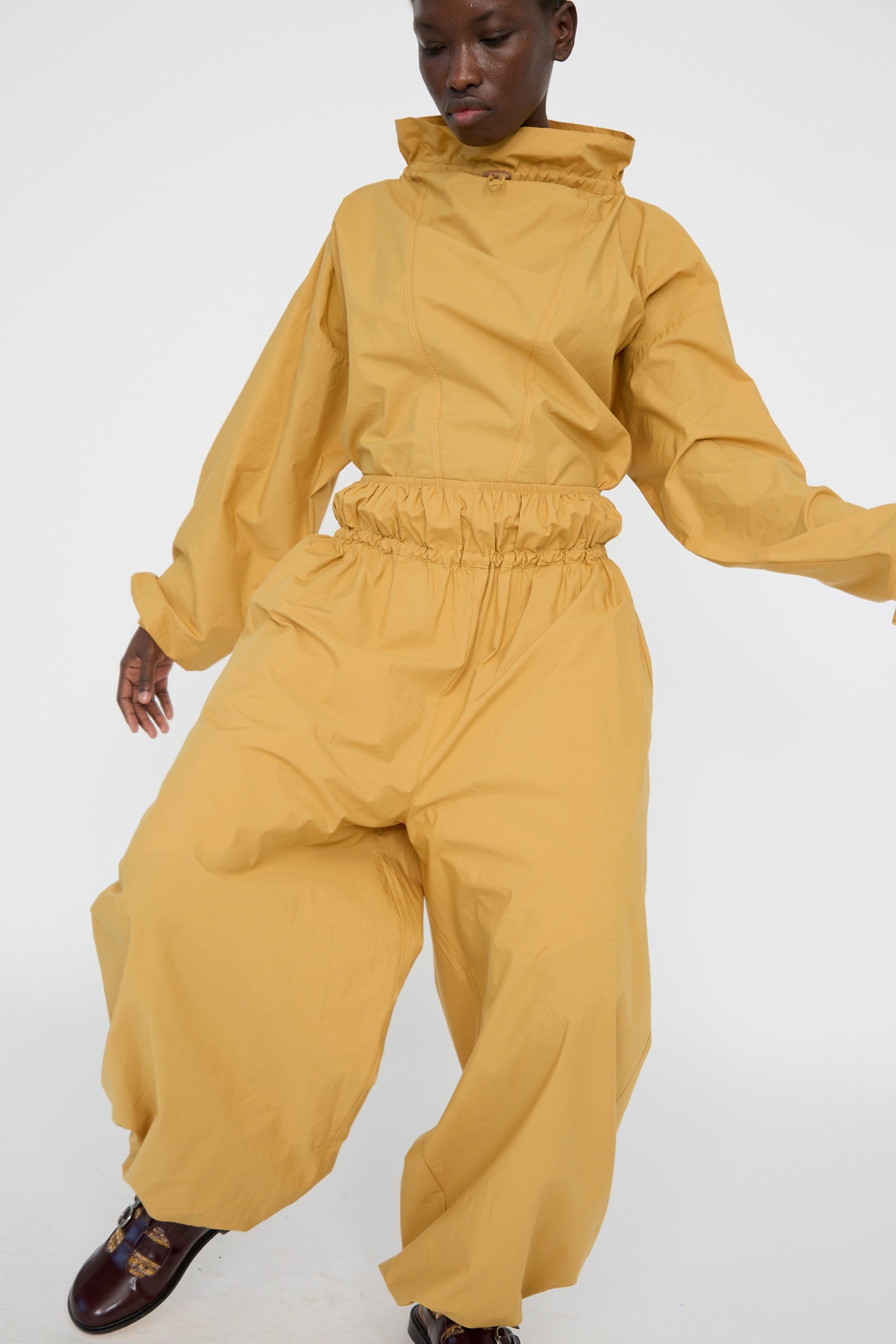 A person models Baserange's Cotton Poplin Air Pants in Sighs Yellow, featuring an oversized and loose-fitting design. Crafted from organic cotton with a high collar, elastic waist, and wide balloon legs, they pose against a plain white background.