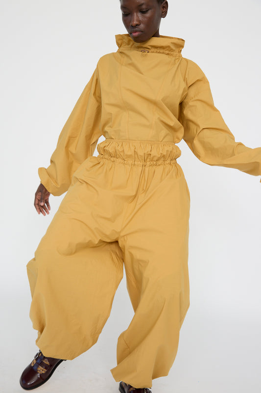 A person models Baserange's Cotton Poplin Air Pants in Sighs Yellow, featuring an oversized and loose-fitting design. Crafted from organic cotton with a high collar, elastic waist, and wide balloon legs, they pose against a plain white background.