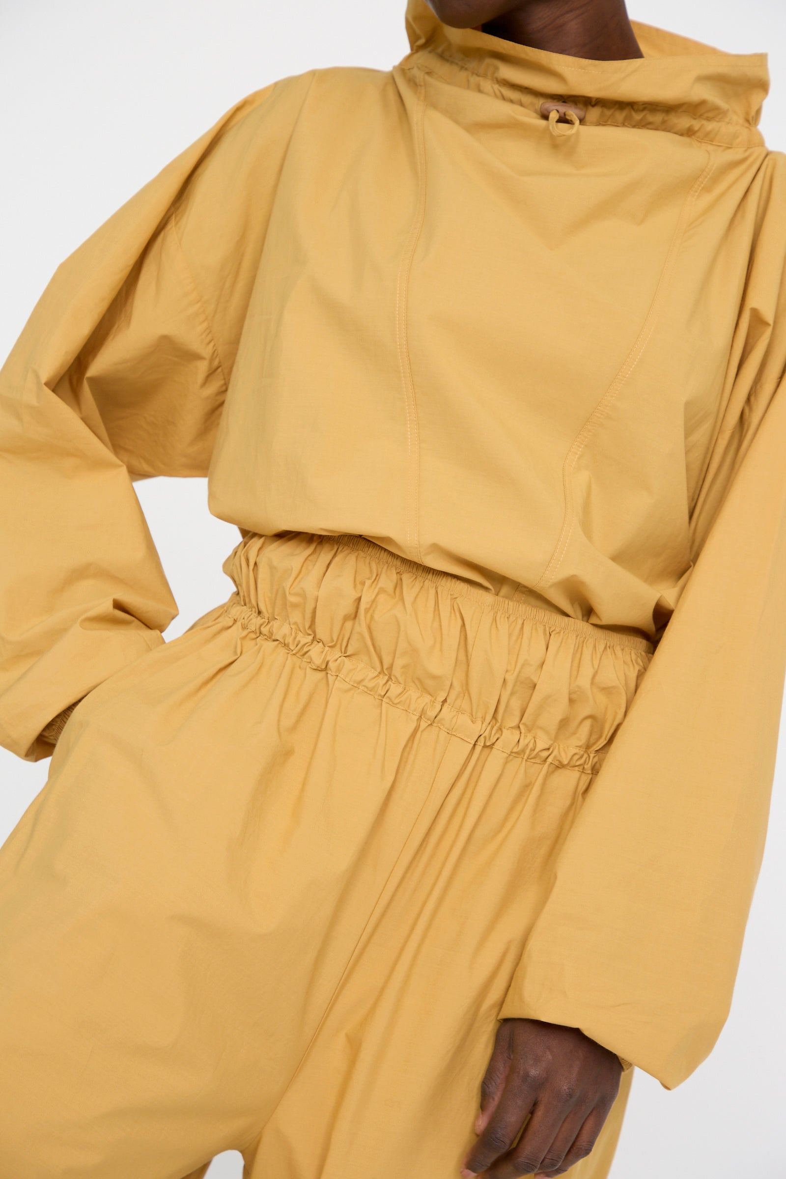 The model is wearing Baserange's Cotton Poplin Air Pants in Sighs Yellow, featuring balloon legs and an elastic waistband, with a hand in the pocket against a white backdrop. 