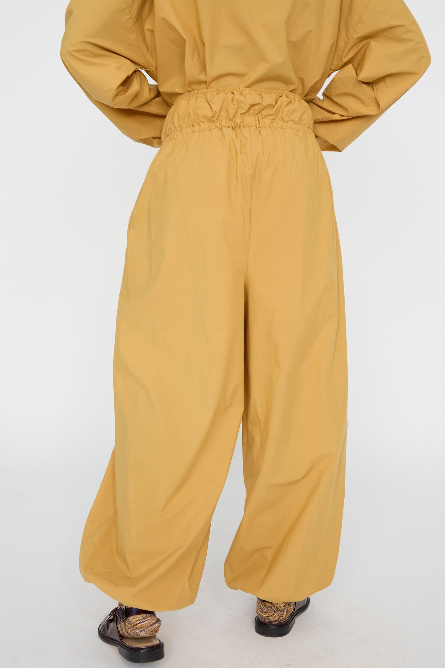 A person is wearing Baserange's Cotton Poplin Air Pants in Sighs Yellow is seen from the back, featuring a loose fit with balloon legs and an elastic waist, made from organic cotton. 