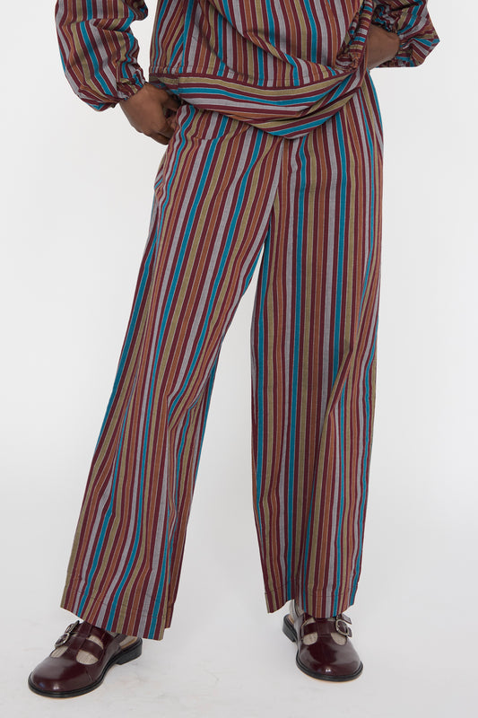 A person is wearing Baserange's matching striped organic cotton outfit with long sleeves and Cotton Poplin Ole Pants in Maroon Multi Stripe, featuring wide-leg and relaxed fit, complemented by shiny burgundy shoes against a white background.