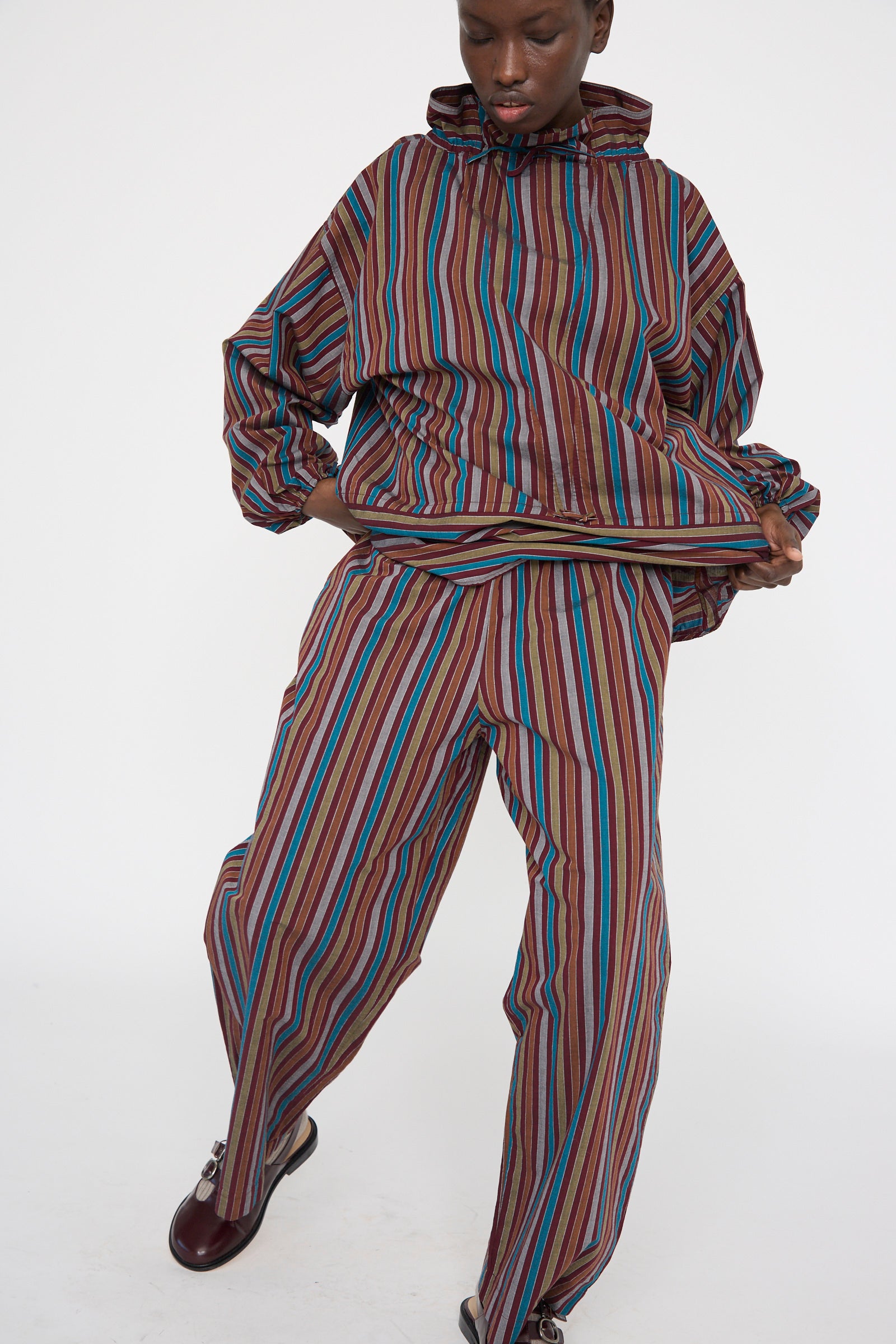 A person stands against a plain white background wearing a colorful striped hooded set and matching relaxed-fit Cotton Poplin Ole Pants in Maroon Multi Stripe by Baserange, crafted from organic cotton.