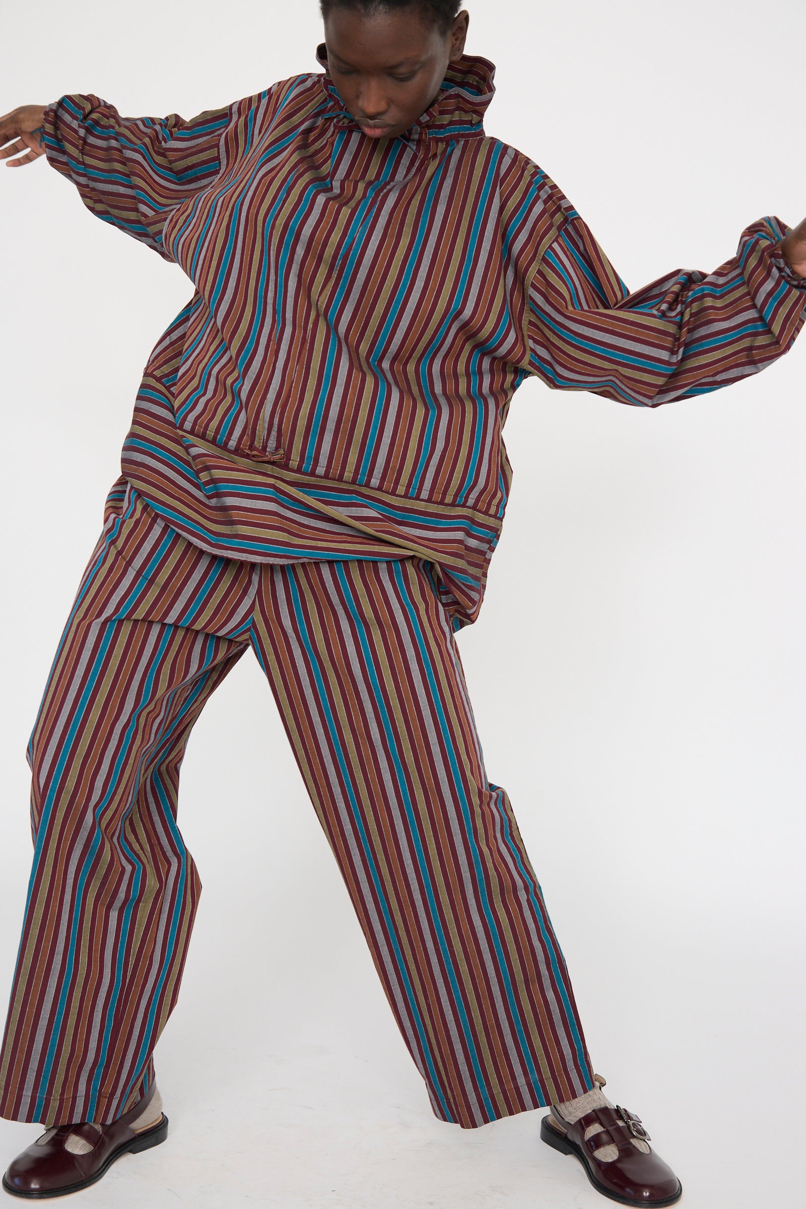 A person in Baserange's Cotton Poplin Ole Pants, featuring maroon multi-stripes, and a matching hooded jacket poses against a white background. Made from organic cotton.