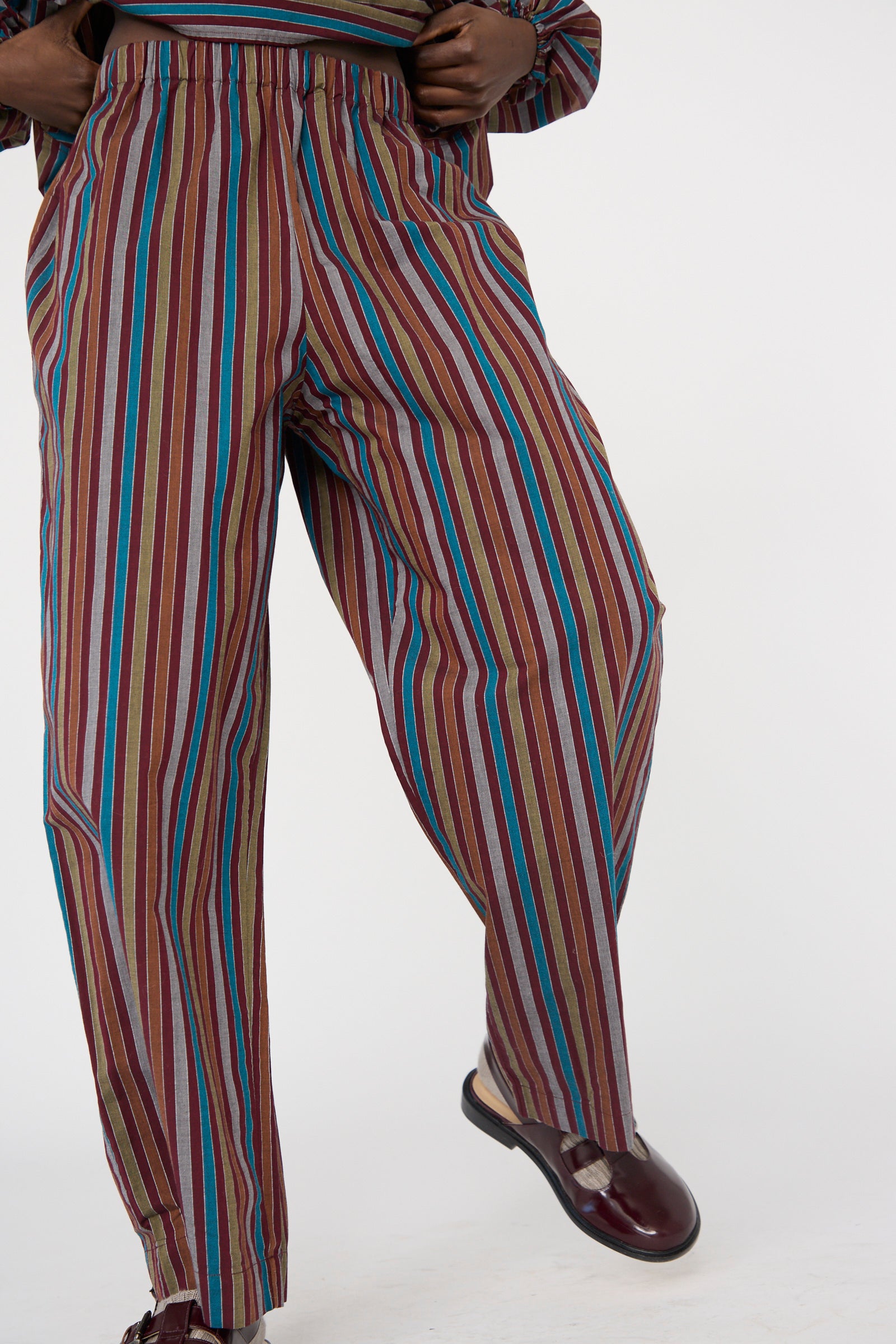 A person models Baserange's Cotton Poplin Ole Pants in Maroon Multi Stripe, featuring a colorful, vertically striped design with wide-leg and relaxed fit, paired with brown loafers against a white background.